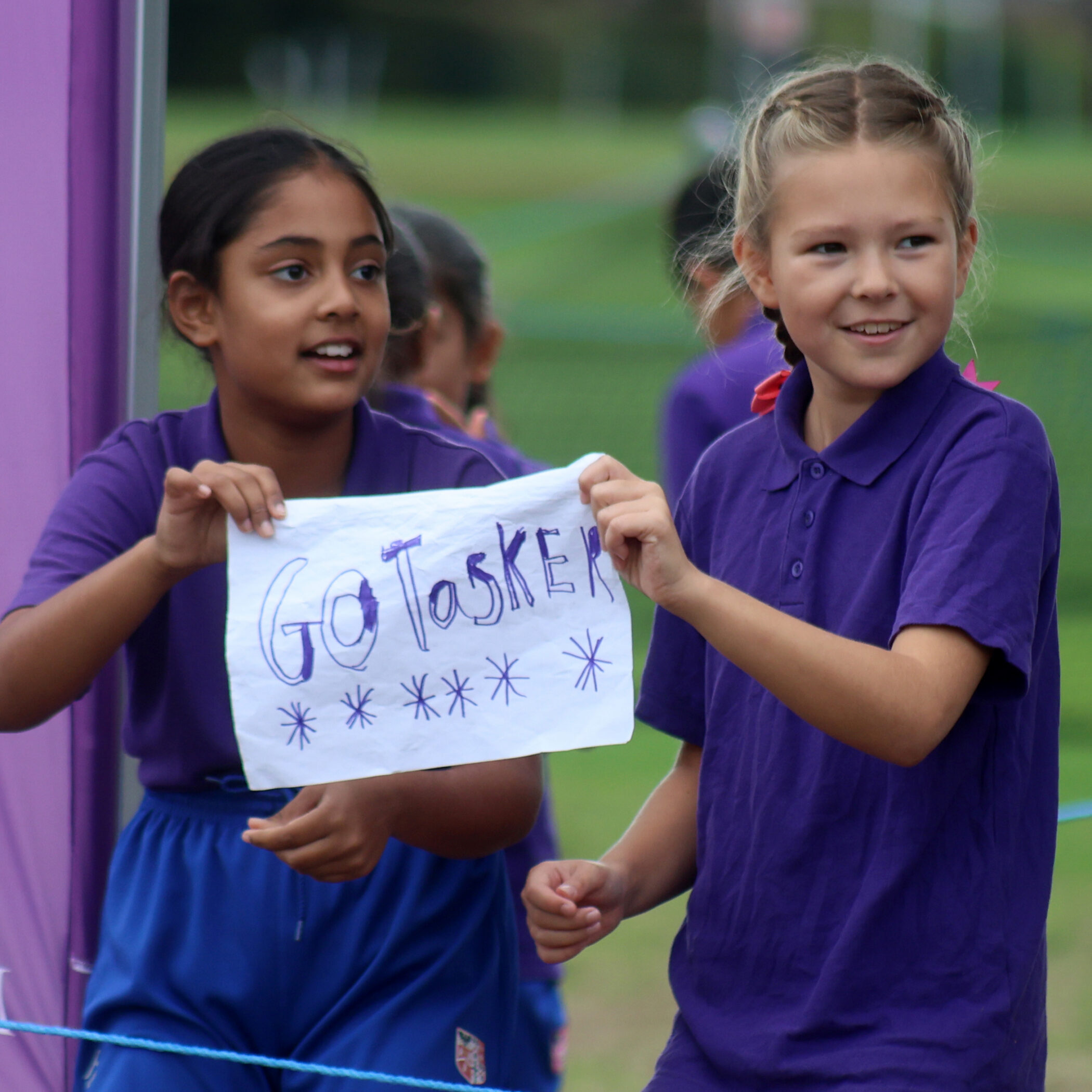 Two girls in purple shirts smile and hold a handmade sign that says Go Tasker with stars, standing outdoors near a purple banner.