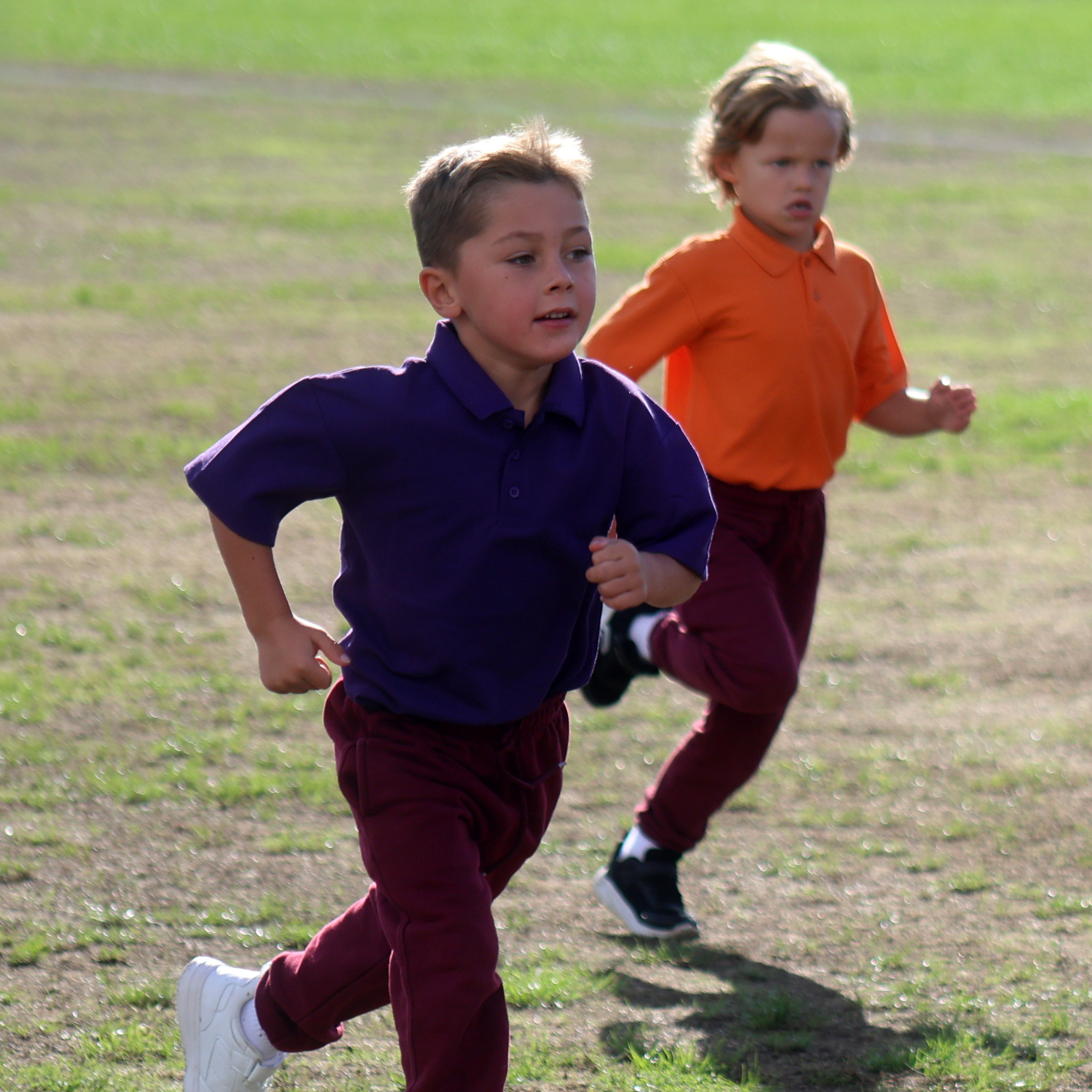 Two young children are running outdoors on a grassy field. The child in front wears a purple shirt and maroon trousers, while the child behind wears an orange shirt and similar maroon trousers.