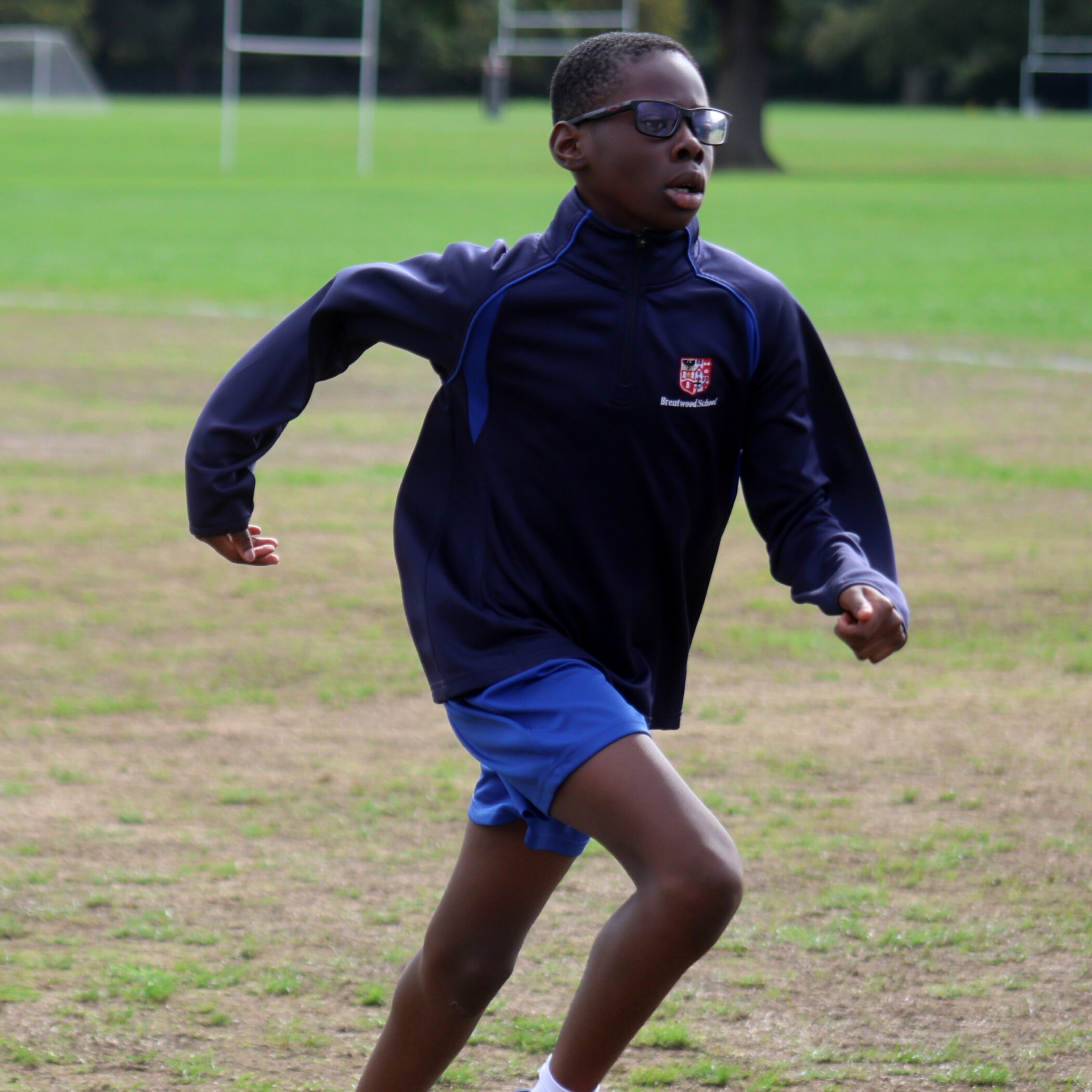 A boy wearing glasses, a navy jacket, and blue shorts is running on a grassy pitch with goalposts in the background.
