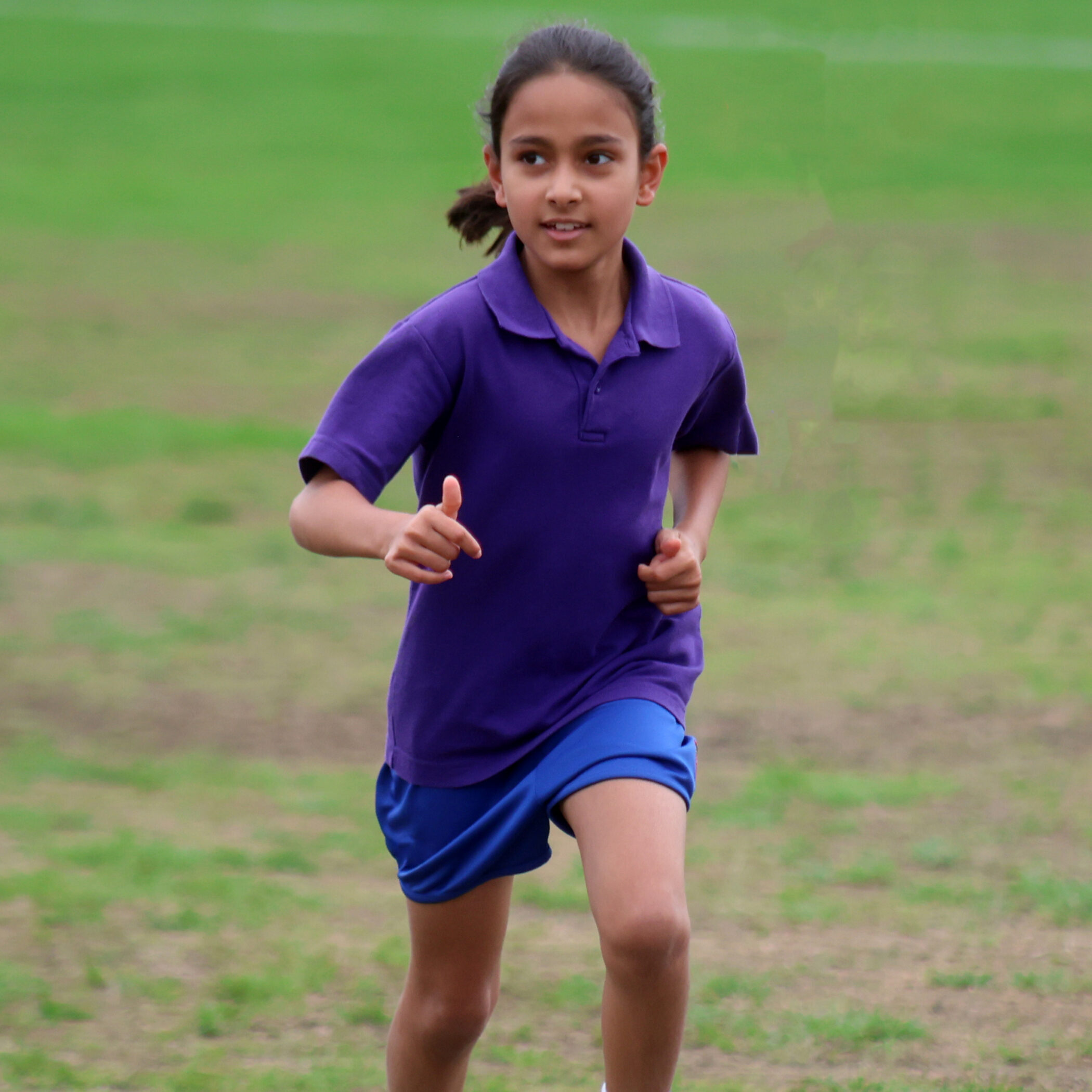 A young girl in a purple shirt and blue shorts is jogging outdoors on a grassy field, looking ahead with a focused expression.