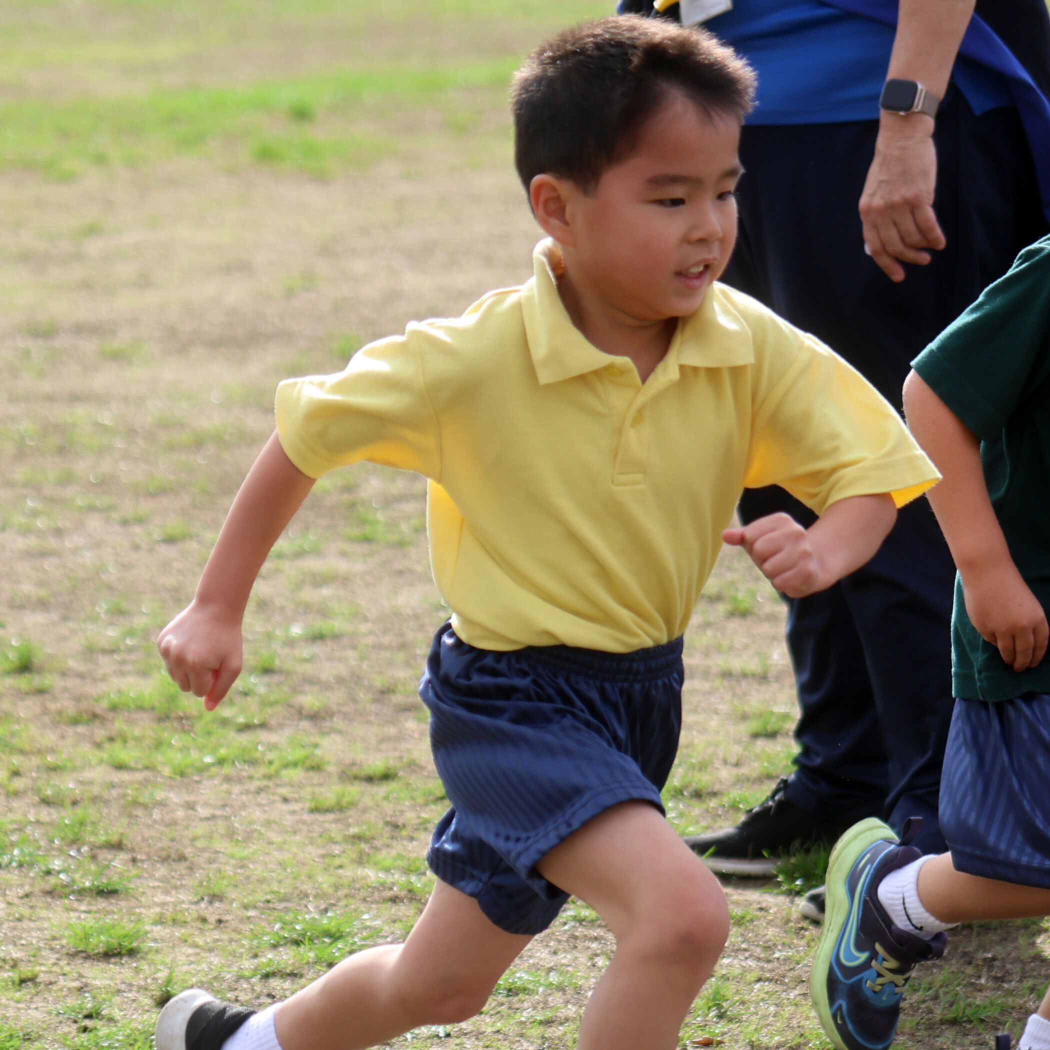 Young boy in a yellow shirt and blue shorts runs outdoors on grass, with another person's hand and leg partially visible beside him.