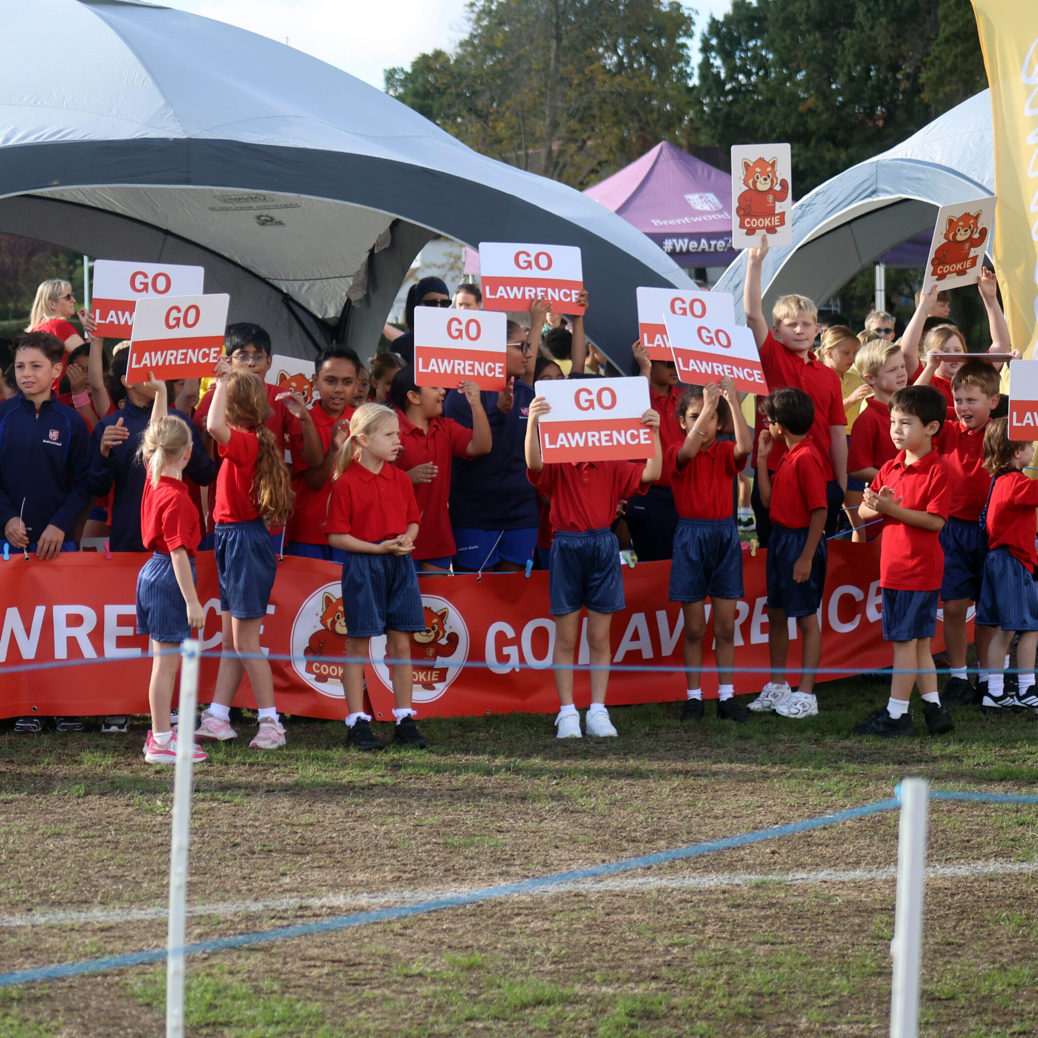 A group of children in red shirts cheer and hold GO LAWRENCE signs behind a red banner at an outdoor sporting event. Some adults and marquees are in the background.
