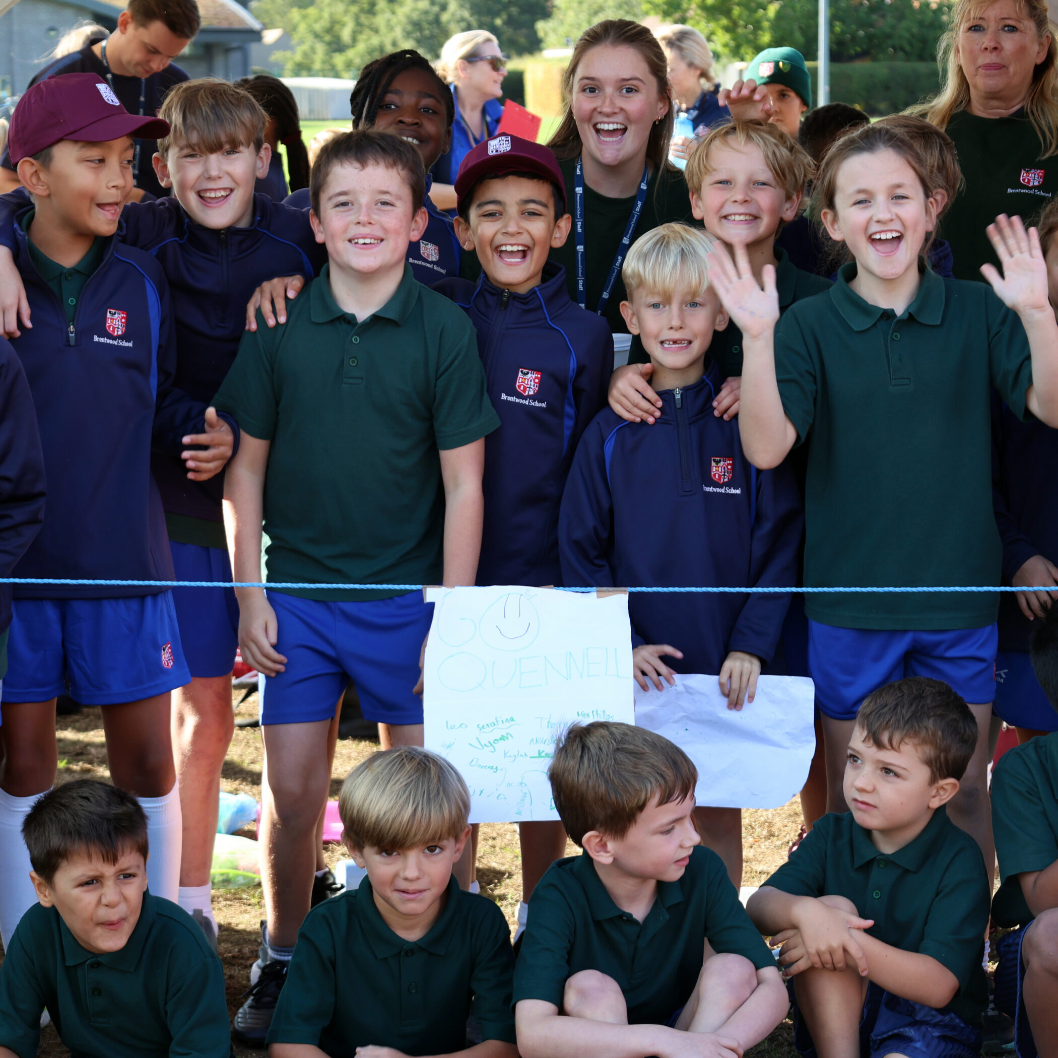 A group of smiling schoolchildren in uniforms pose outdoors, some standing and some sitting. One child holds a homemade sign, and several children are waving at the camera.