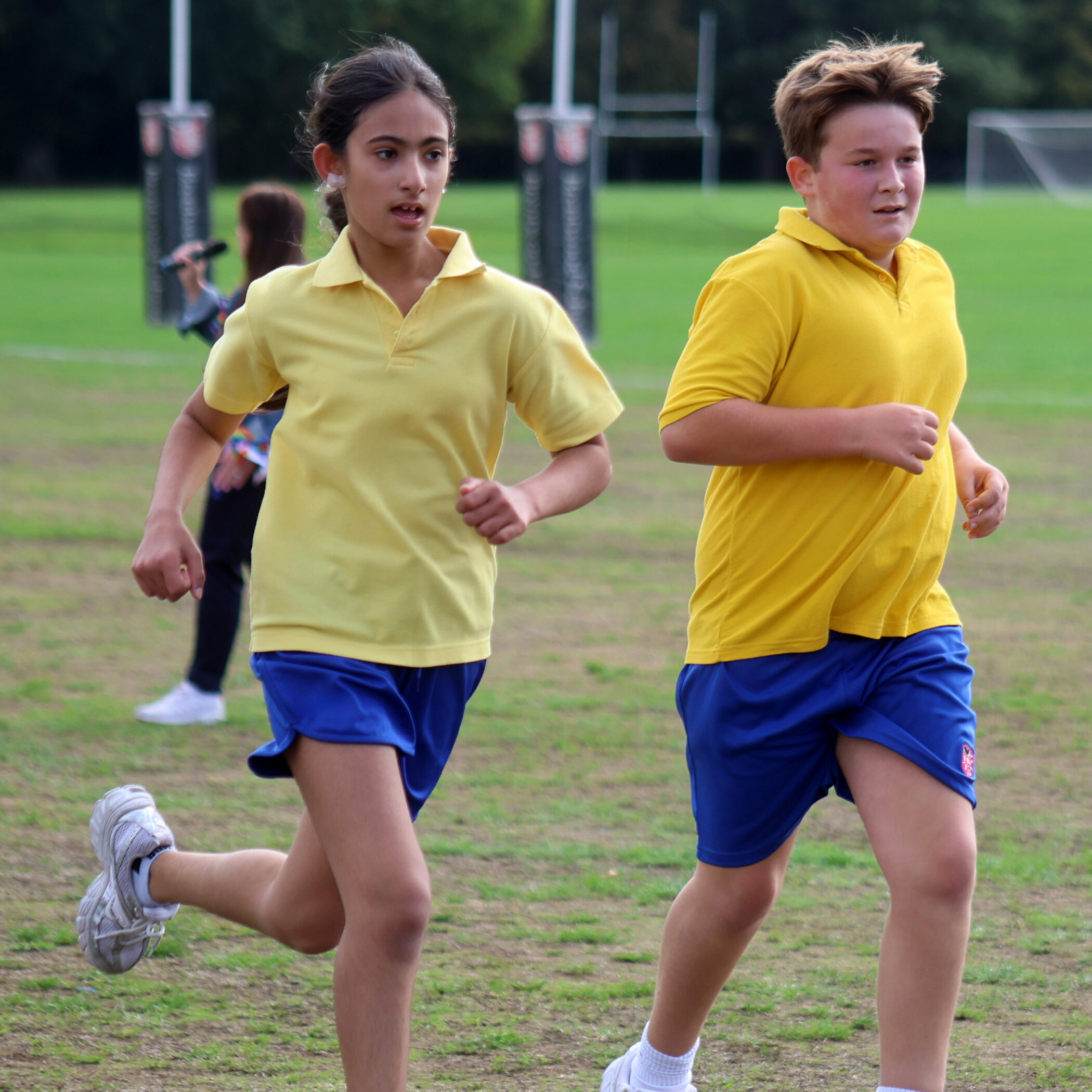 Two children wearing yellow shirts and blue shorts are running on a grassy sports pitch. In the background, goalposts and another person can be seen. Both children appear focused and are participating in an outdoor activity.