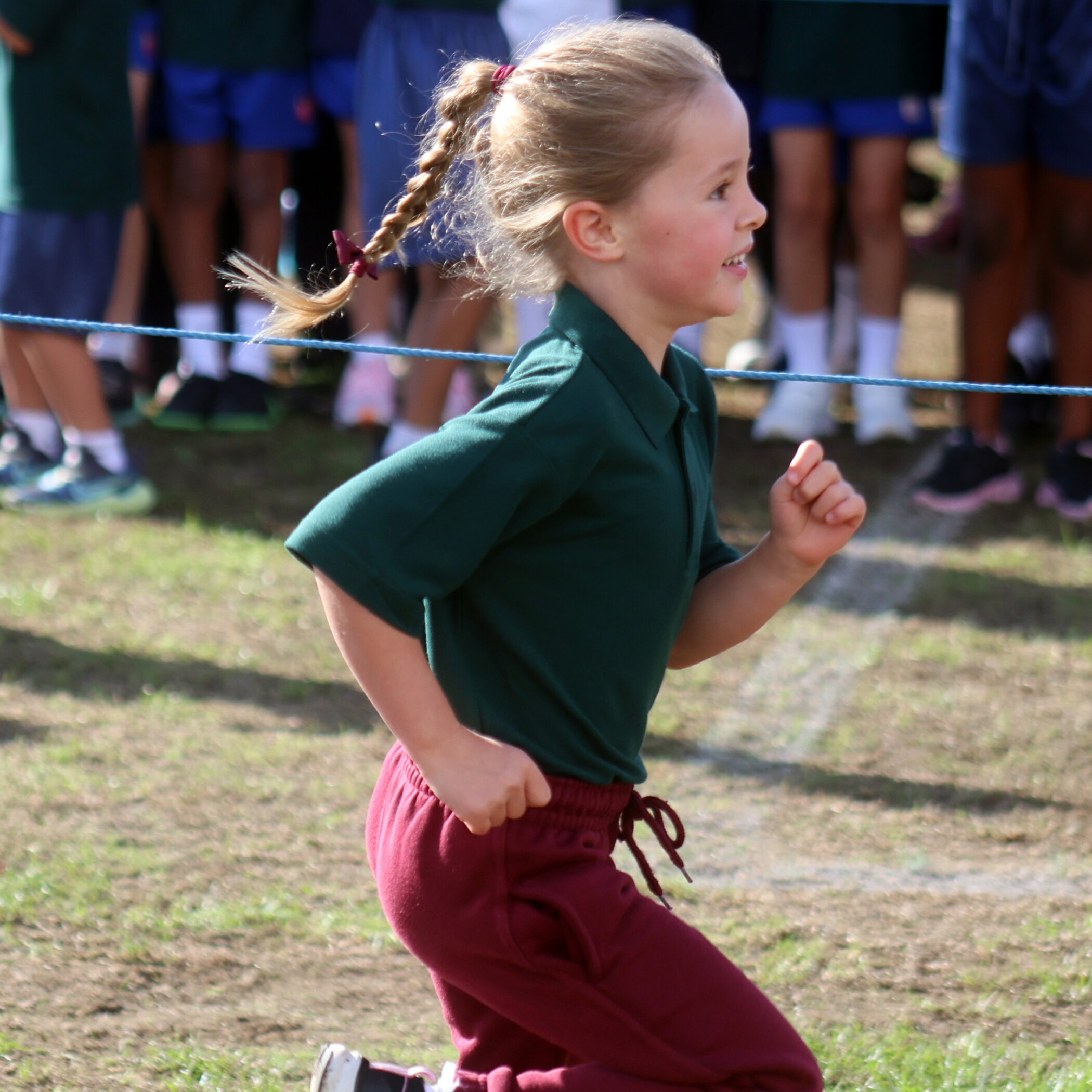 A young child in a green shirt and red trousers runs outside during a race, with a group of people watching in the background.