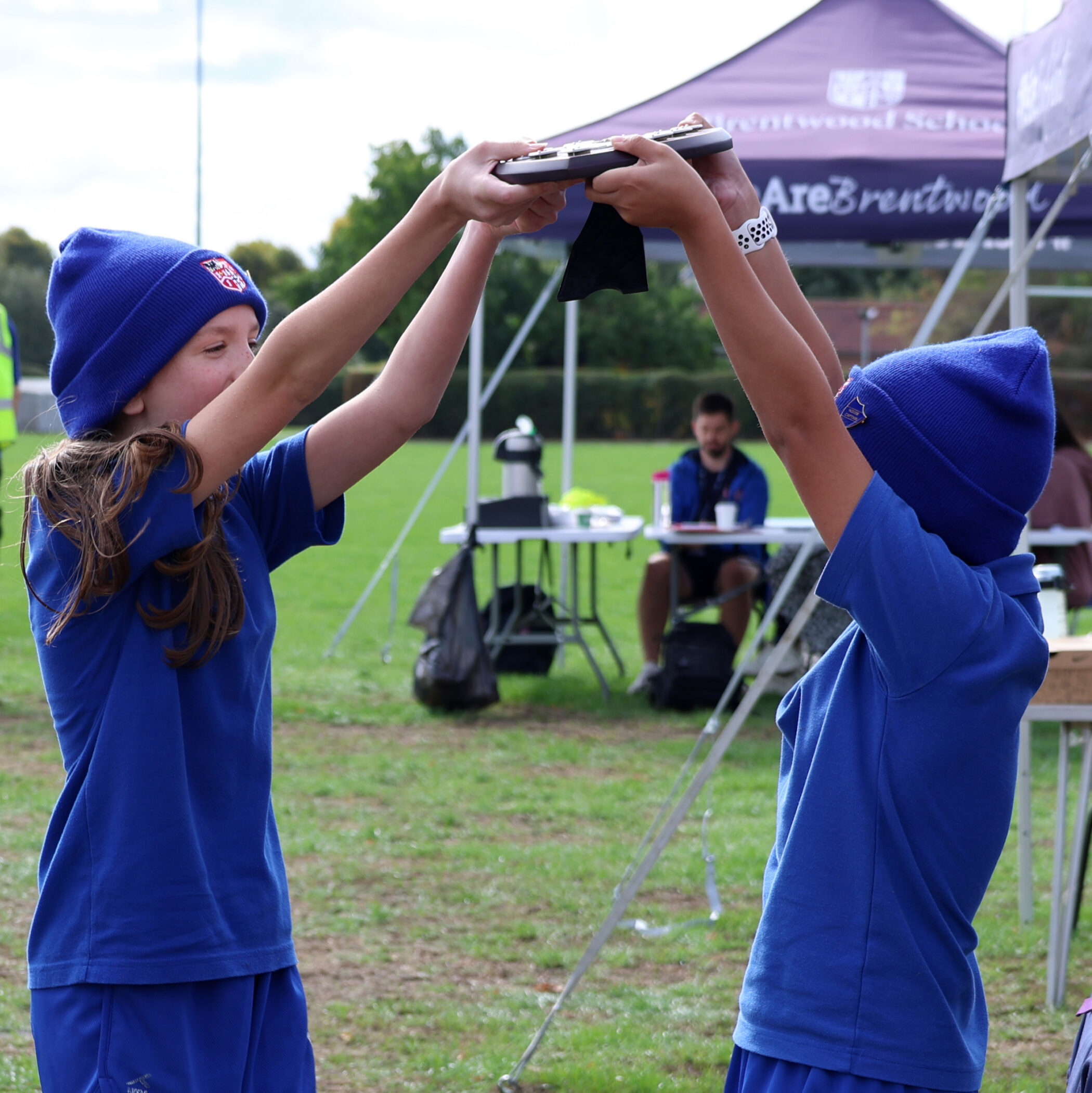 Two children wearing blue uniforms and hats lift a trophy together outdoors, smiling. A man sits at a table in the background under a marquee, with green grass and trees around.