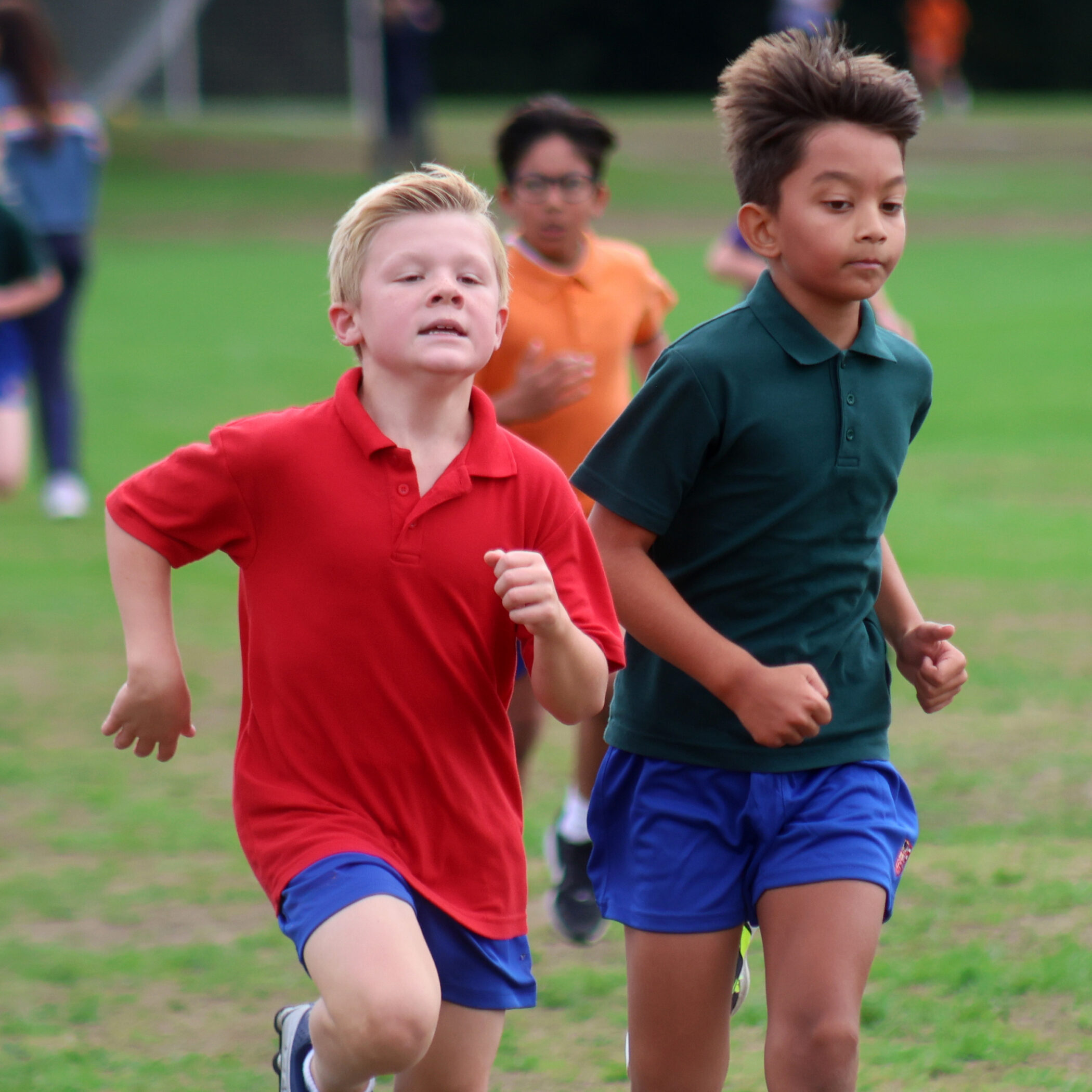 Two boys in sports uniforms race on a grassy field, focused and determined, with other children running behind them during what appears to be a school sports event.