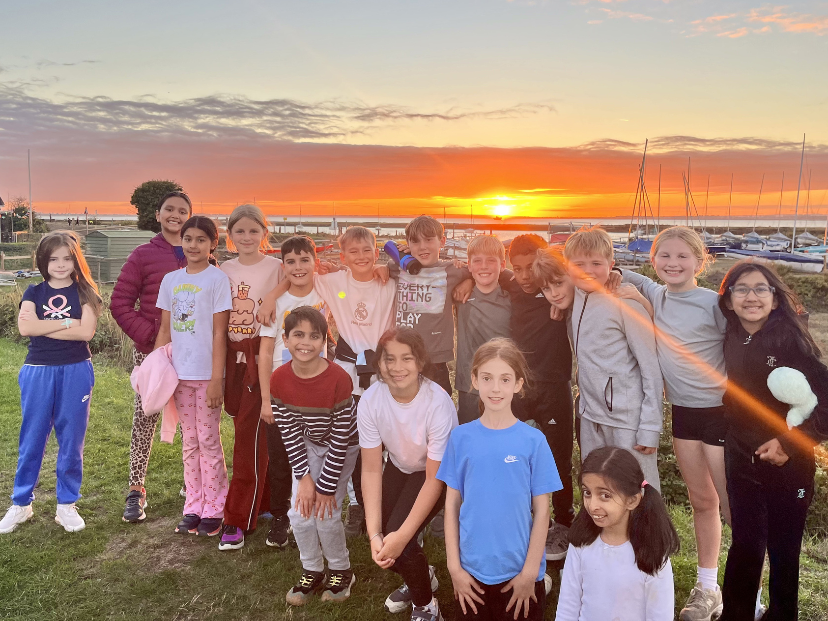 A group of children pose and smile together outdoors at sunset, with sailing boats and the sea in the background. They appear happy and relaxed, enjoying a bright, colourful evening.