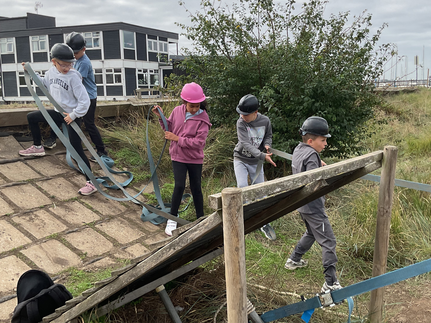 Five children wearing helmets are outdoors on a wooden assault course, pulling on straps and climbing a sloped platform, with grassy terrain and a building in the background.