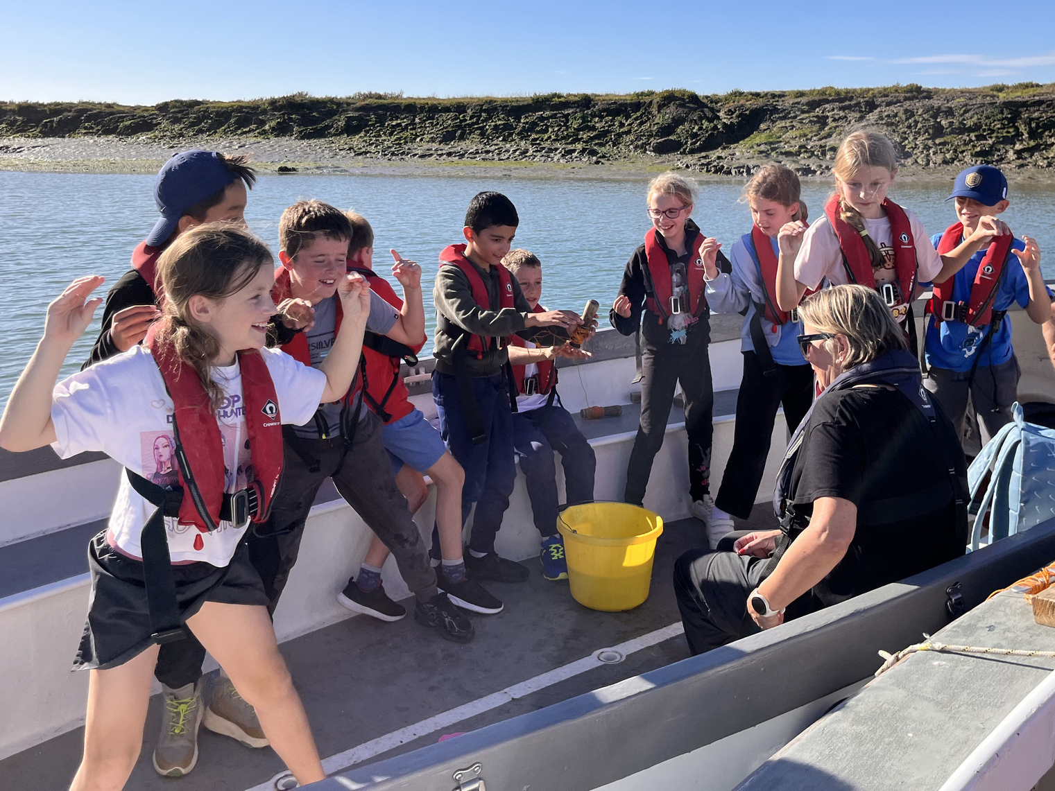 A group of children in life jackets excitedly react to something in a yellow bucket whilst sitting in a boat with an adult on a sunny day by the water.