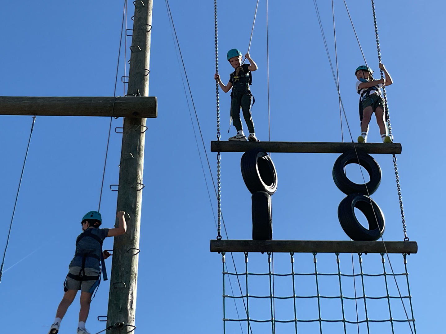 Three children wearing helmets and harnesses climb a high ropes course with wooden beams, tyres, and nets, set against a clear blue sky.