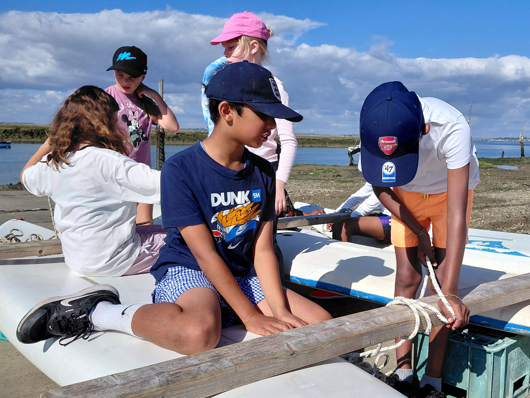 Five children sit and work on small boats by the water, tying ropes and wearing casual summer clothes and caps, with blue sky and clouds in the background.
