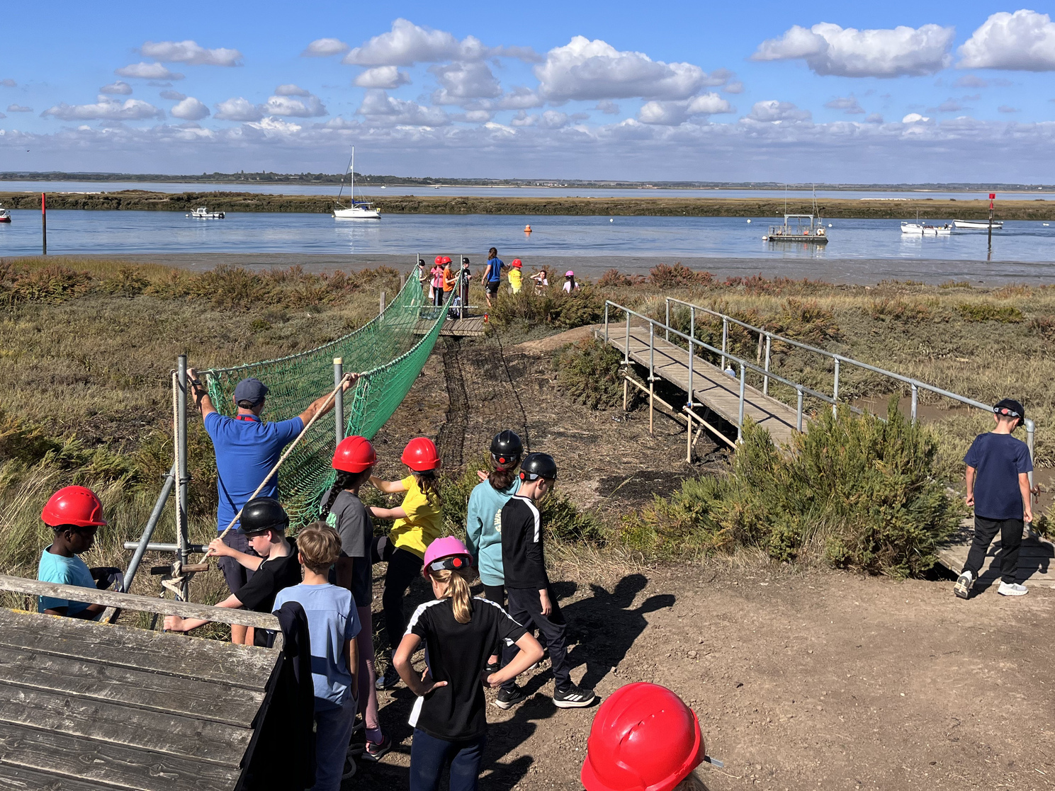 Children in helmets queue for a zipwire across a marshy area near water, with boats and people in the background under a partly cloudy sky.