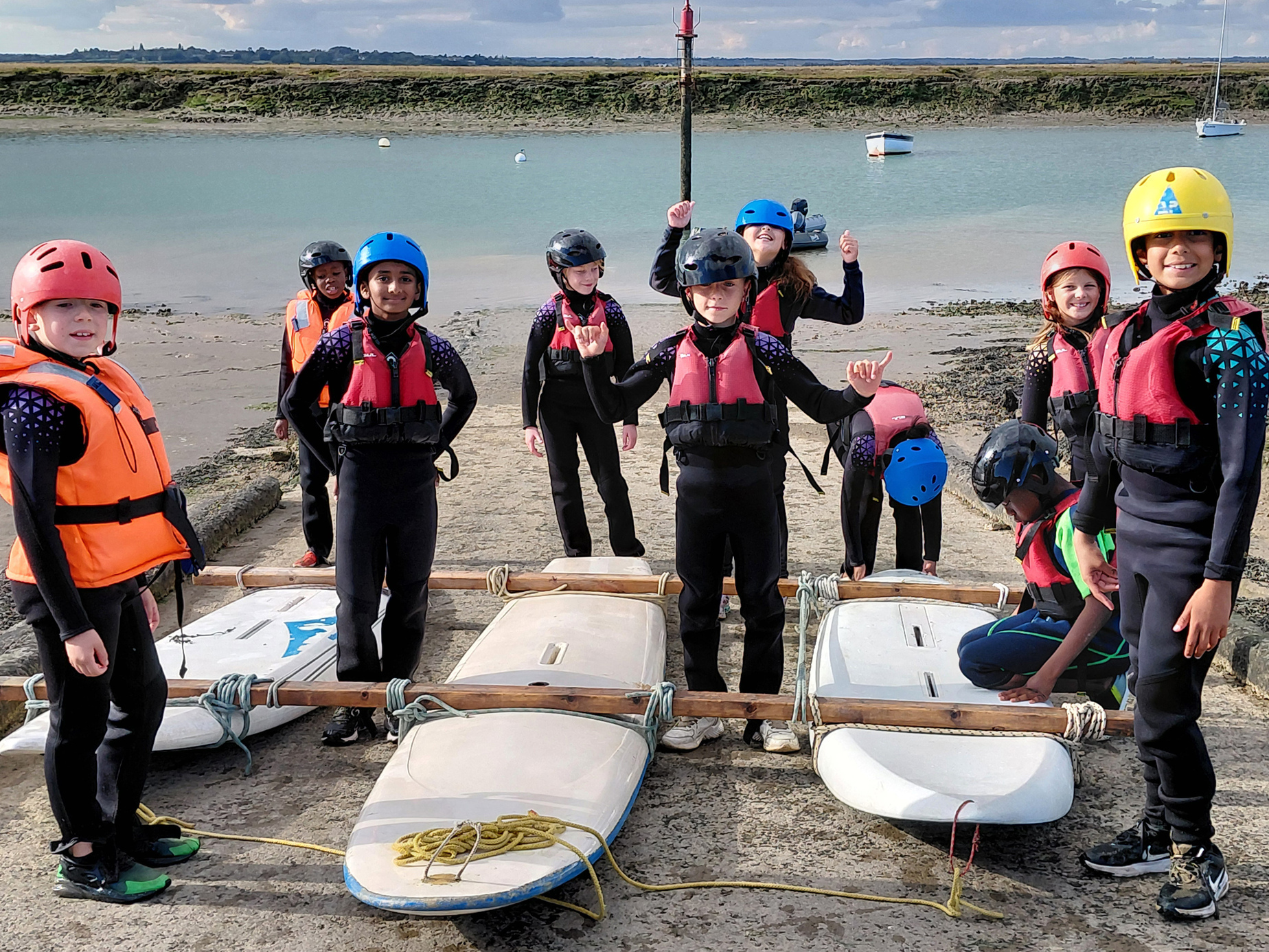 A group of children wearing helmets, life jackets, and wetsuits stand on a jetty by surfboards, smiling and posing before water activities on a cloudy day.