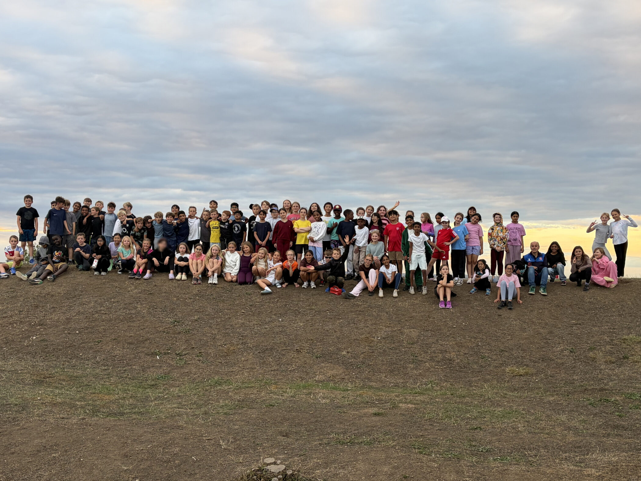 A large group of children and adults pose together on a grassy hill under a cloudy sky, with some sitting and others standing.