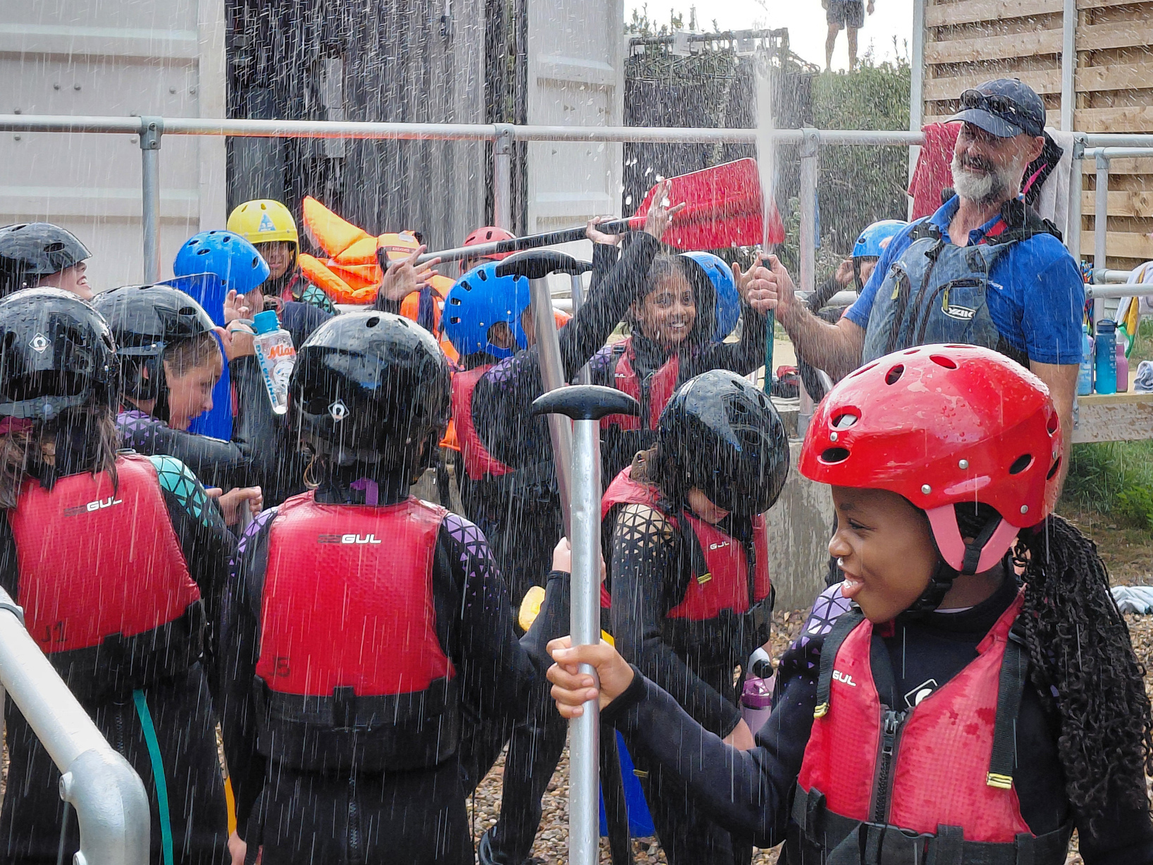 A group of people in helmets and life jackets laugh under a spray of water outdoors, some holding paddles. An adult, also in gear, holds the hose, and everyone appears to be having fun.