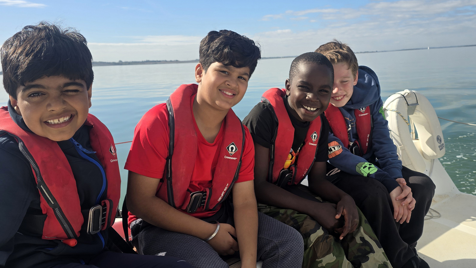 Four boys wearing red life jackets sit side by side on a boat, smiling at the camera. Calm water and blue sky are visible in the background.