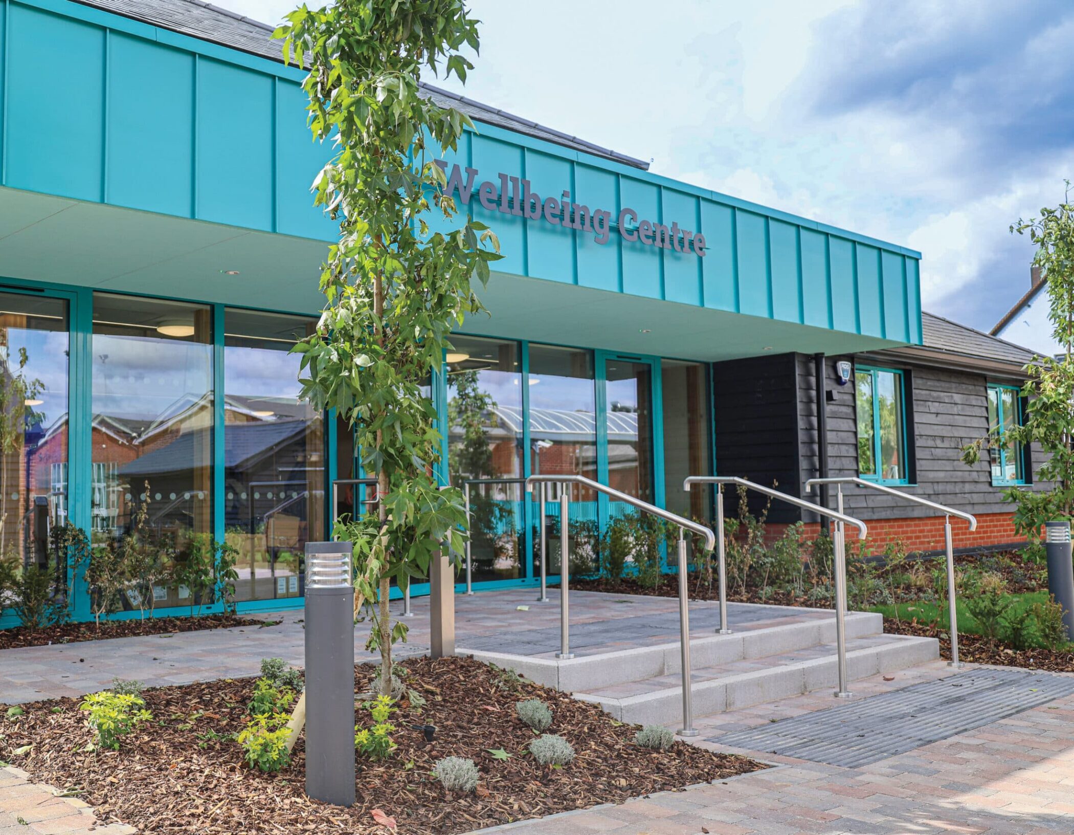 A modern building with teal panels and glass doors, labelled Wellbeing Centre, stands as the focal point of the Wellbeing Centre Development. Steps, railings, greenery, and landscaped gardens are visible in the foreground under partly cloudy skies.