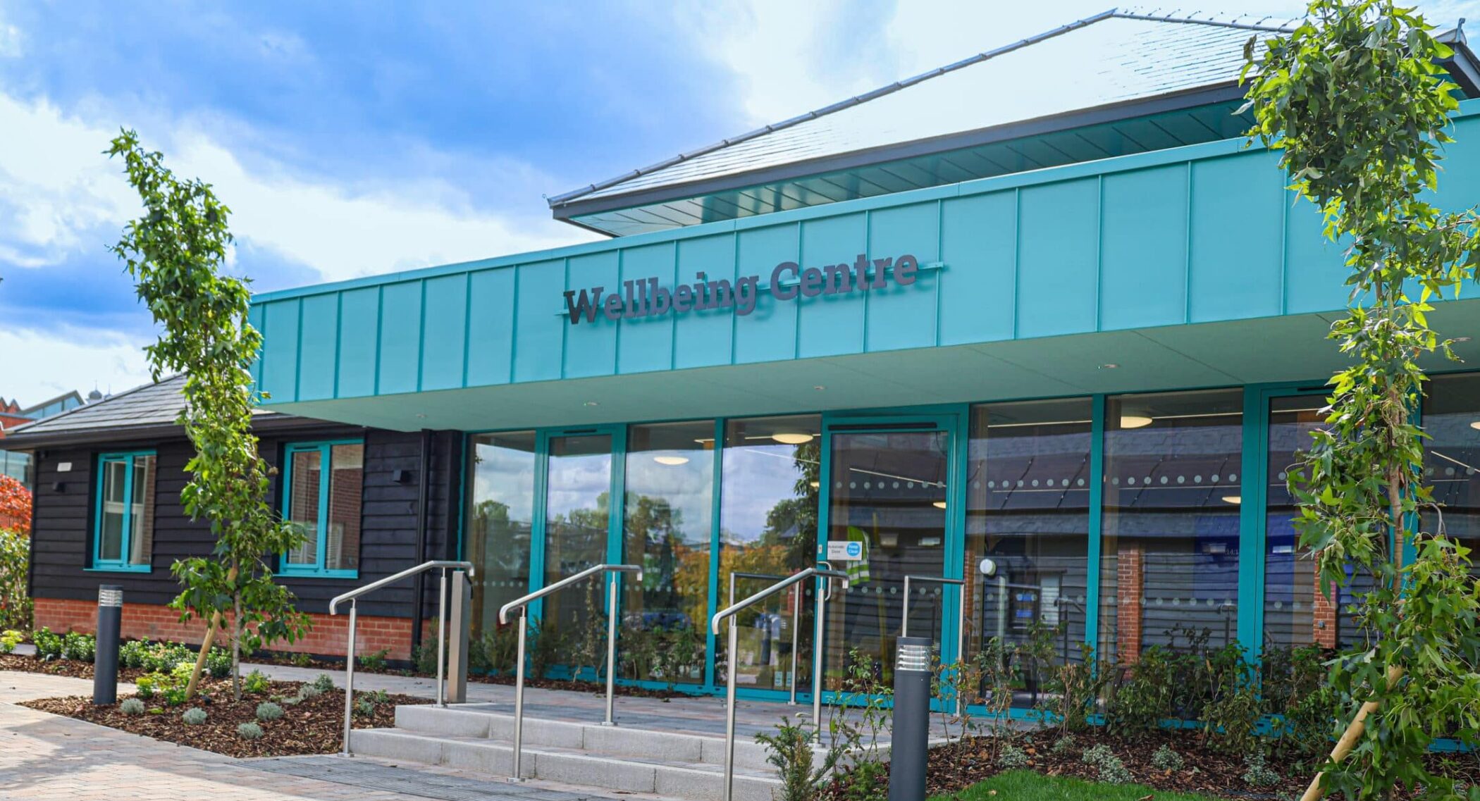 A modern building labelled Wellbeing Centre, part of an exciting Wellbeing Centre Development, features large glass doors, teal accents, a brick walkway, benches, and young trees in front, all set under a partly cloudy sky.