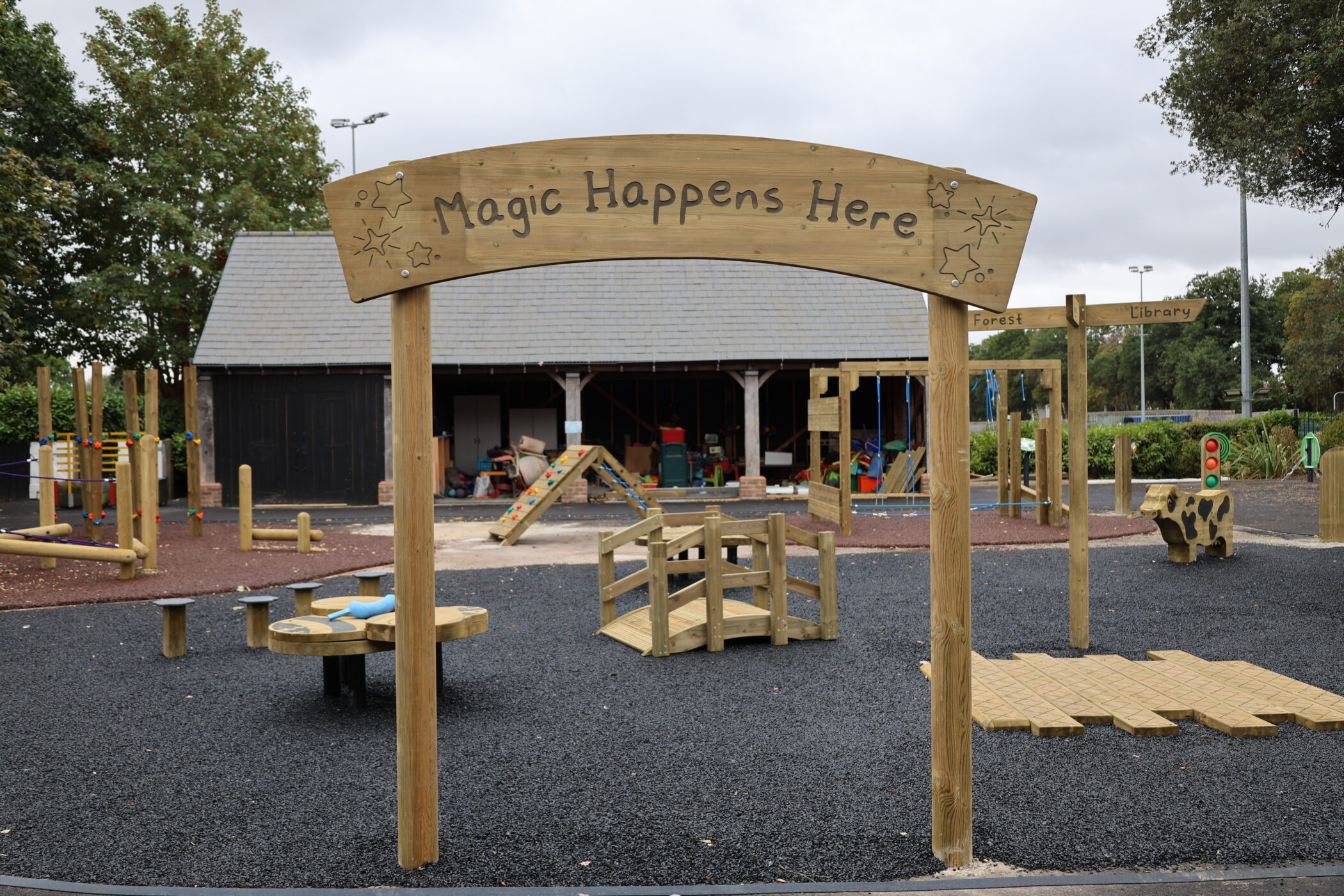 A playground with a wooden arch reading Magic Happens Here, play equipment, and a small building in the background. The area features various facilities, including wooden signs, and is surrounded by trees.