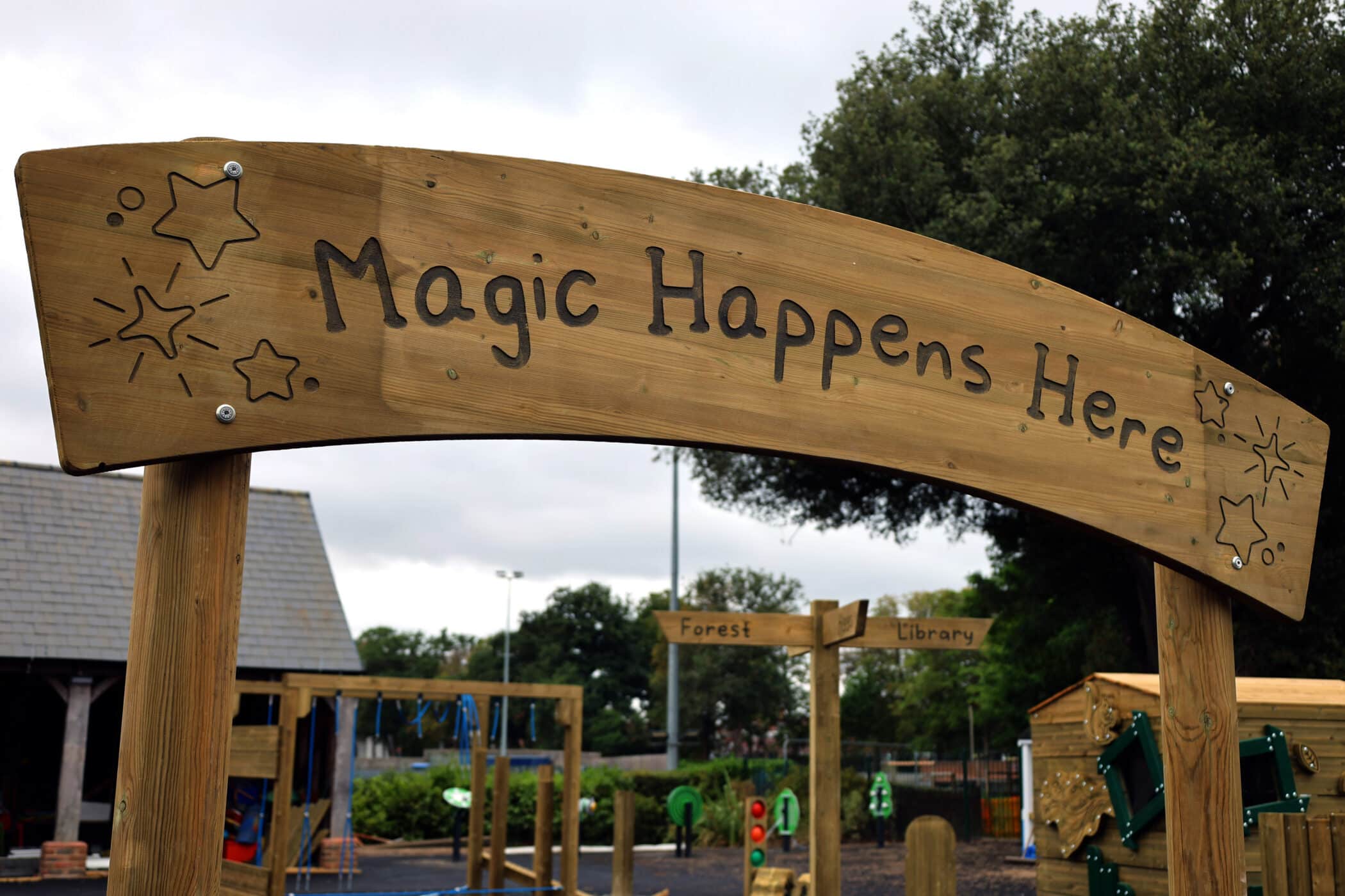 A wooden sign decorated with stars reads Magic Happens Here at the entrance to a playground, with play structures and trees visible in the background.