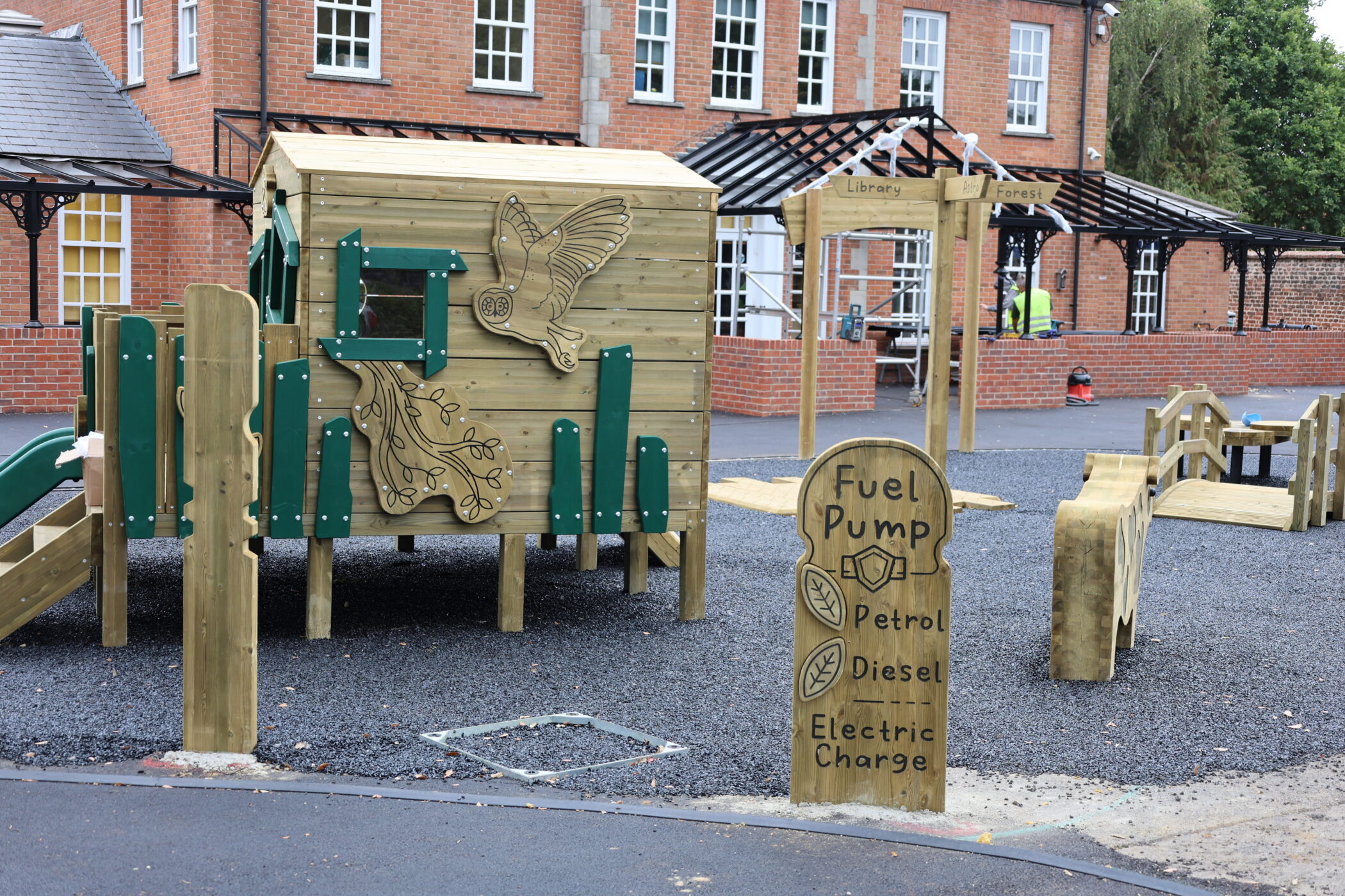 A wooden playground with animal carvings, a slide, bridge, and a whimsical Fuel Pump sign listing petrol, diesel, and electric charge is set in front of a red brick building with white windows, highlighting the creative facilities designed for imaginative play.