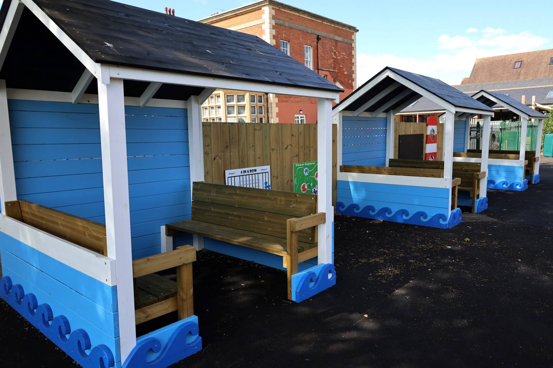 Three blue wooden beach hut-style shelters with benches are lined up on a paved outdoor area, with a brick building and fence in the background under a clear sky.