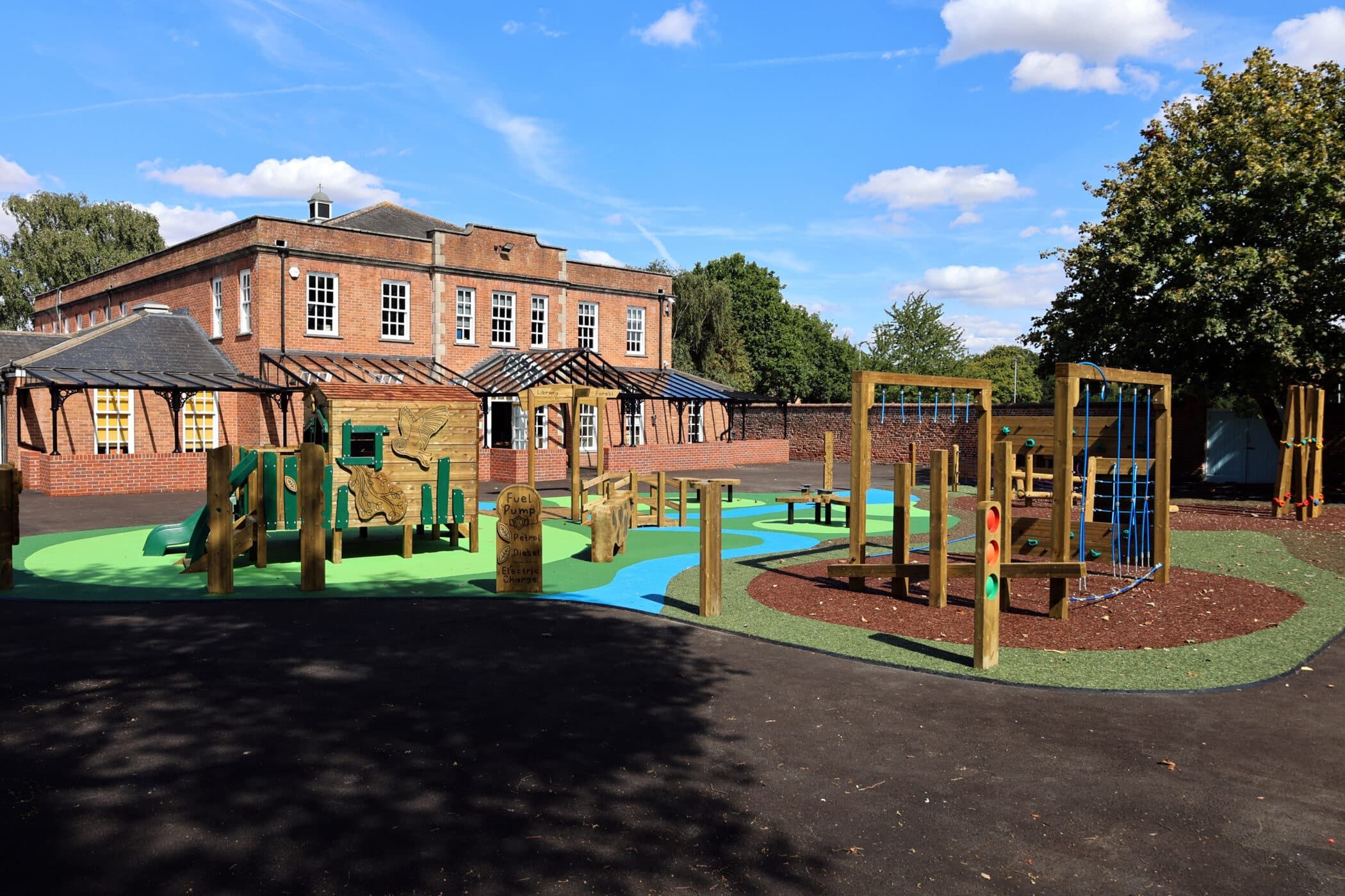 A colourful playground with climbing frames and slides sits in front of a large brick building under a blue sky with scattered clouds. Trees border the play area.