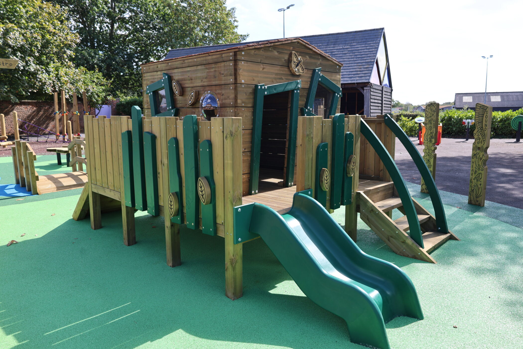 Wooden playground structure with green double slide, climbing steps, and a ramp, set on a green rubber surface. The well-maintained facilities are surrounded by trees and buildings, all visible in the background under a sunny sky.