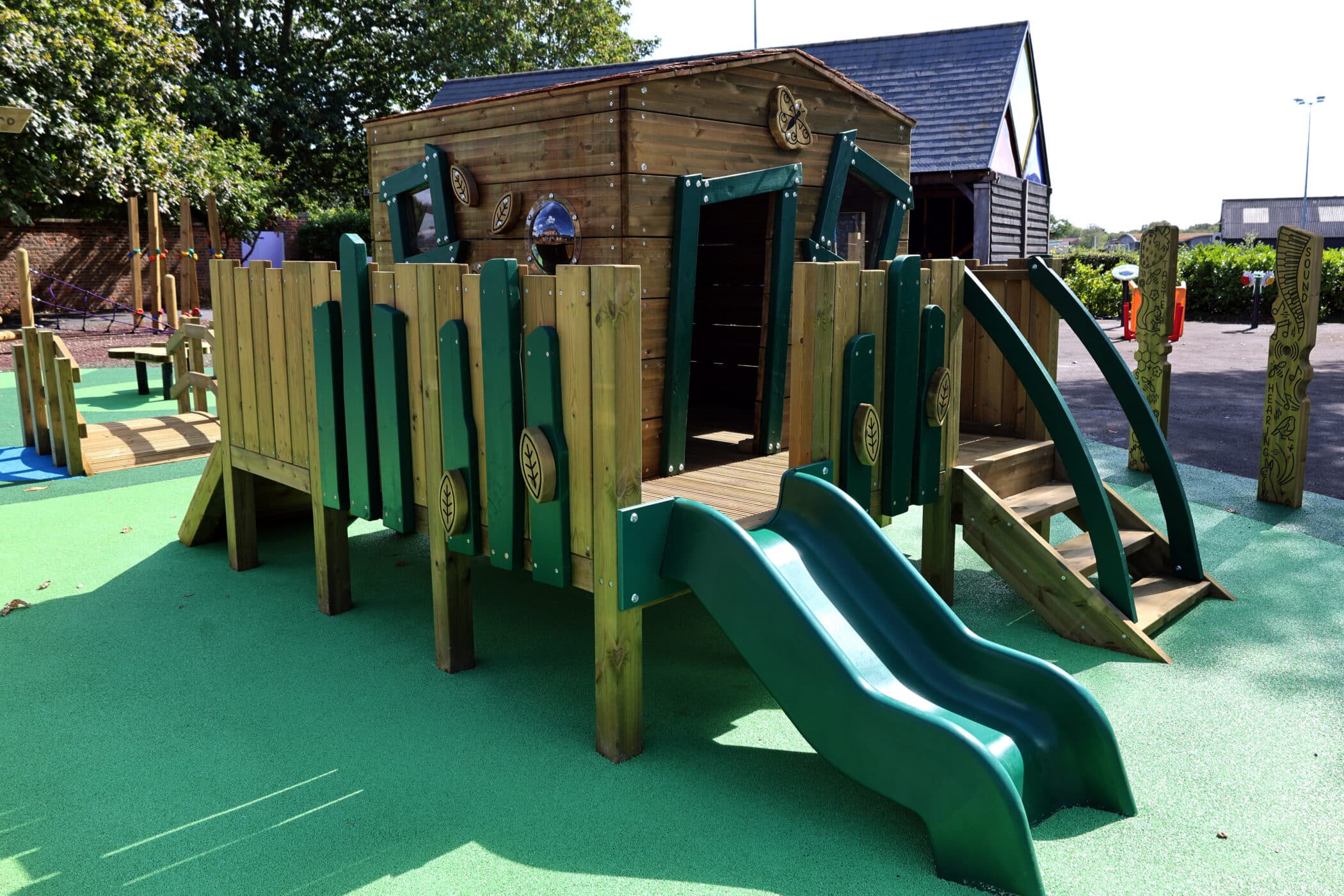 Wooden playground structure with two green slides, small climbing ramps, and a bridge set on green safety flooring. There is a wooden playhouse structure on top and trees in the background.
