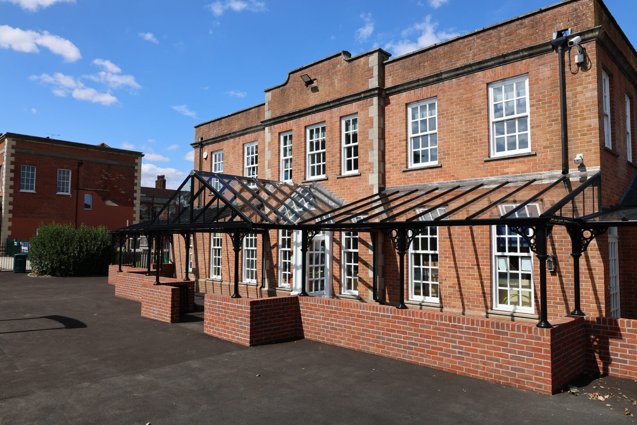 A red-brick building with tall windows and a glass-roofed walkway, set against a blue sky with scattered clouds. The area in front is paved and spacious, providing easy access to the building’s modern facilities.