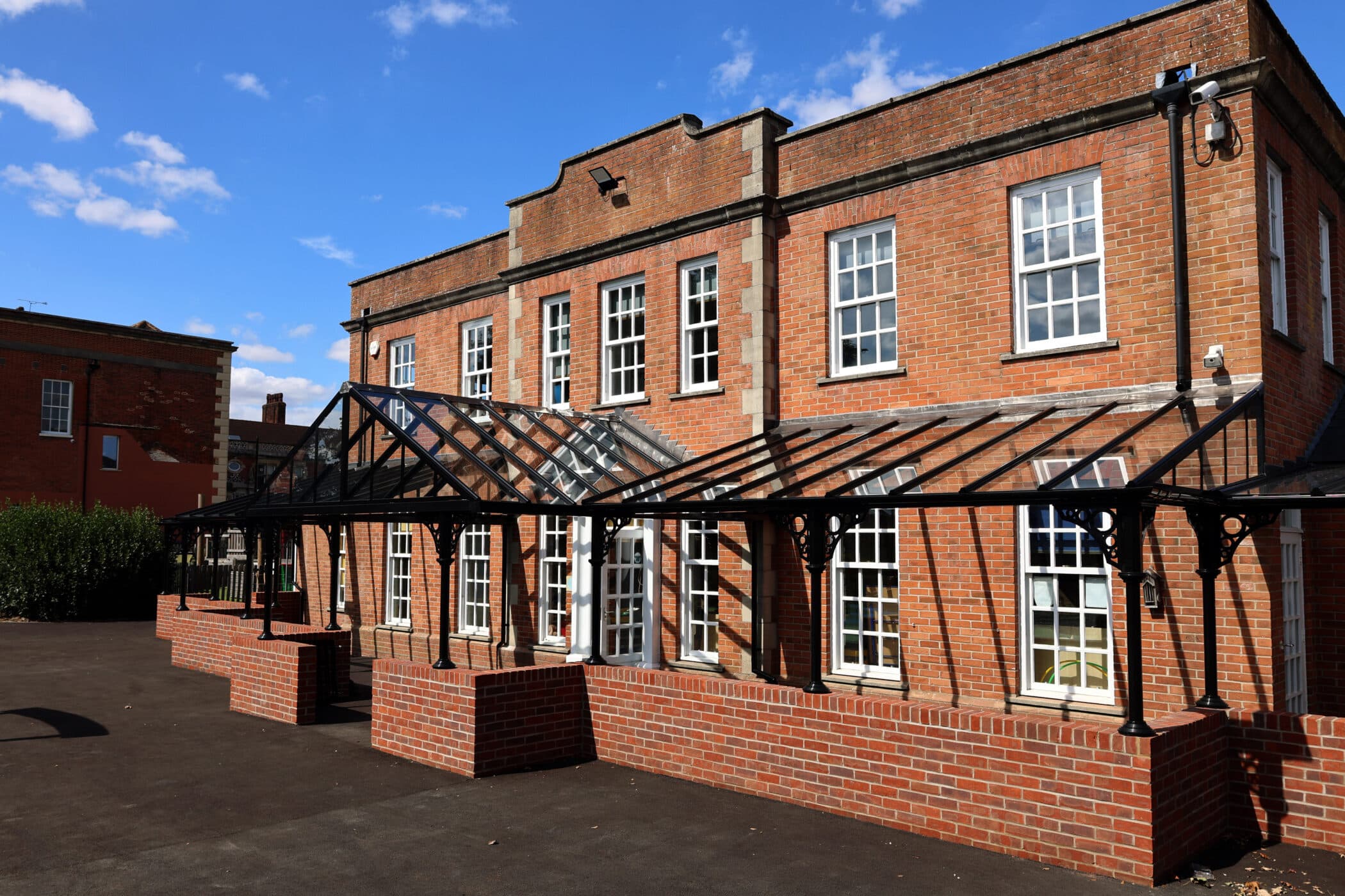 A red brick building with white-framed windows and a glass-and-metal canopy over the entrance, set against a blue sky with scattered clouds.