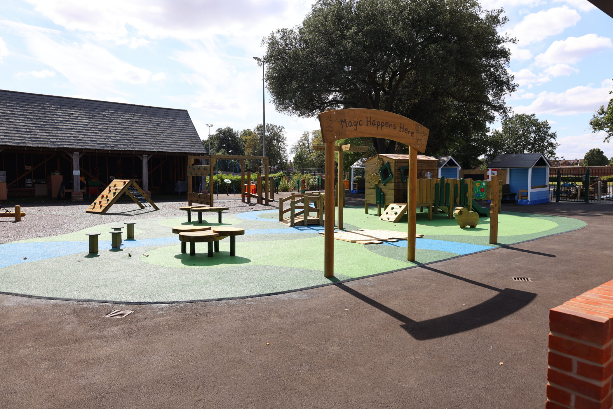 A colourful outdoor playground with climbing structures and well-maintained facilities, featuring benches and a sign reading Magic Happens Here under a large tree, with buildings and greenery in the background.