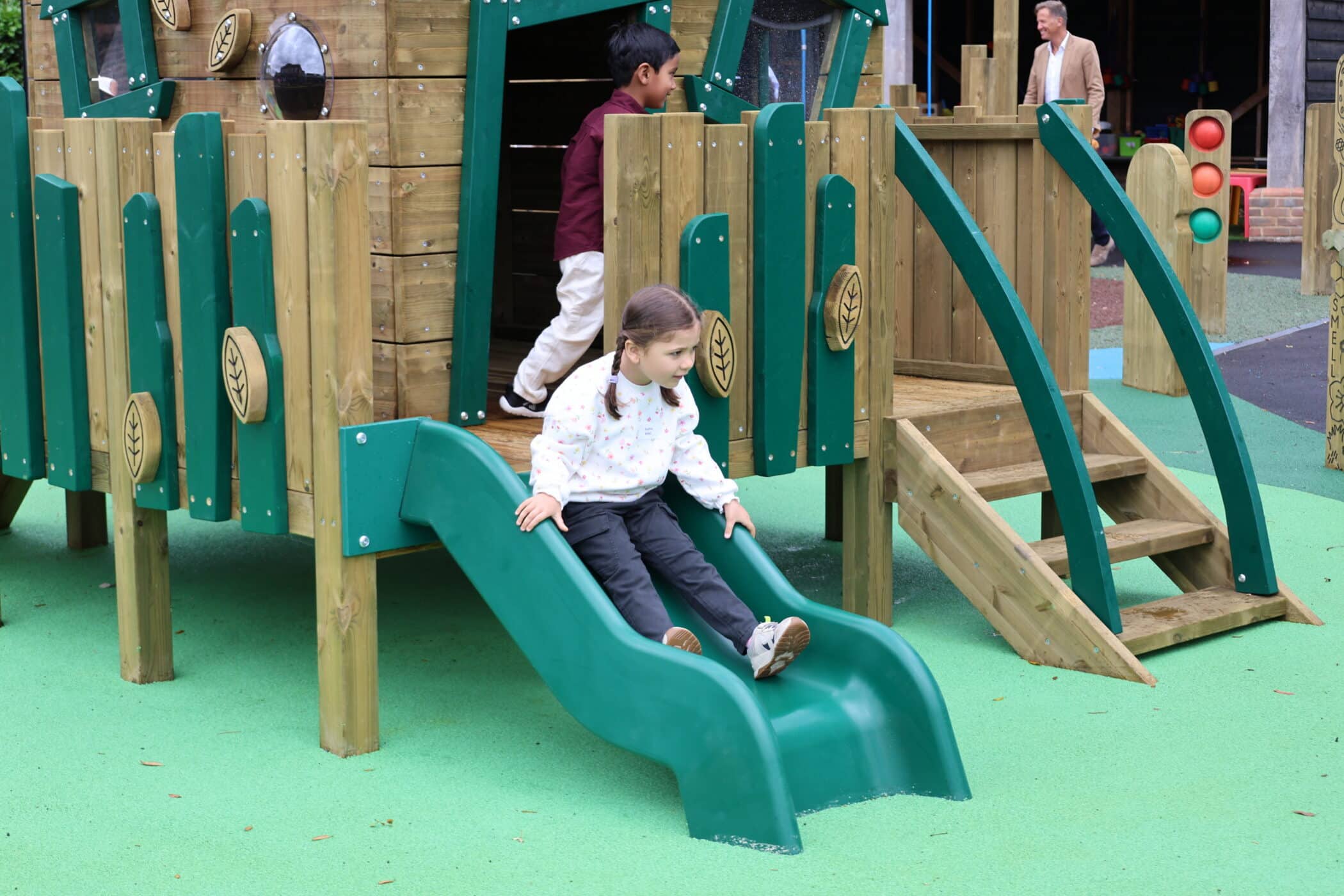 A young girl slides down a green slide attached to a wooden playground structure whilst a boy stands at the top. An adult is in the background.