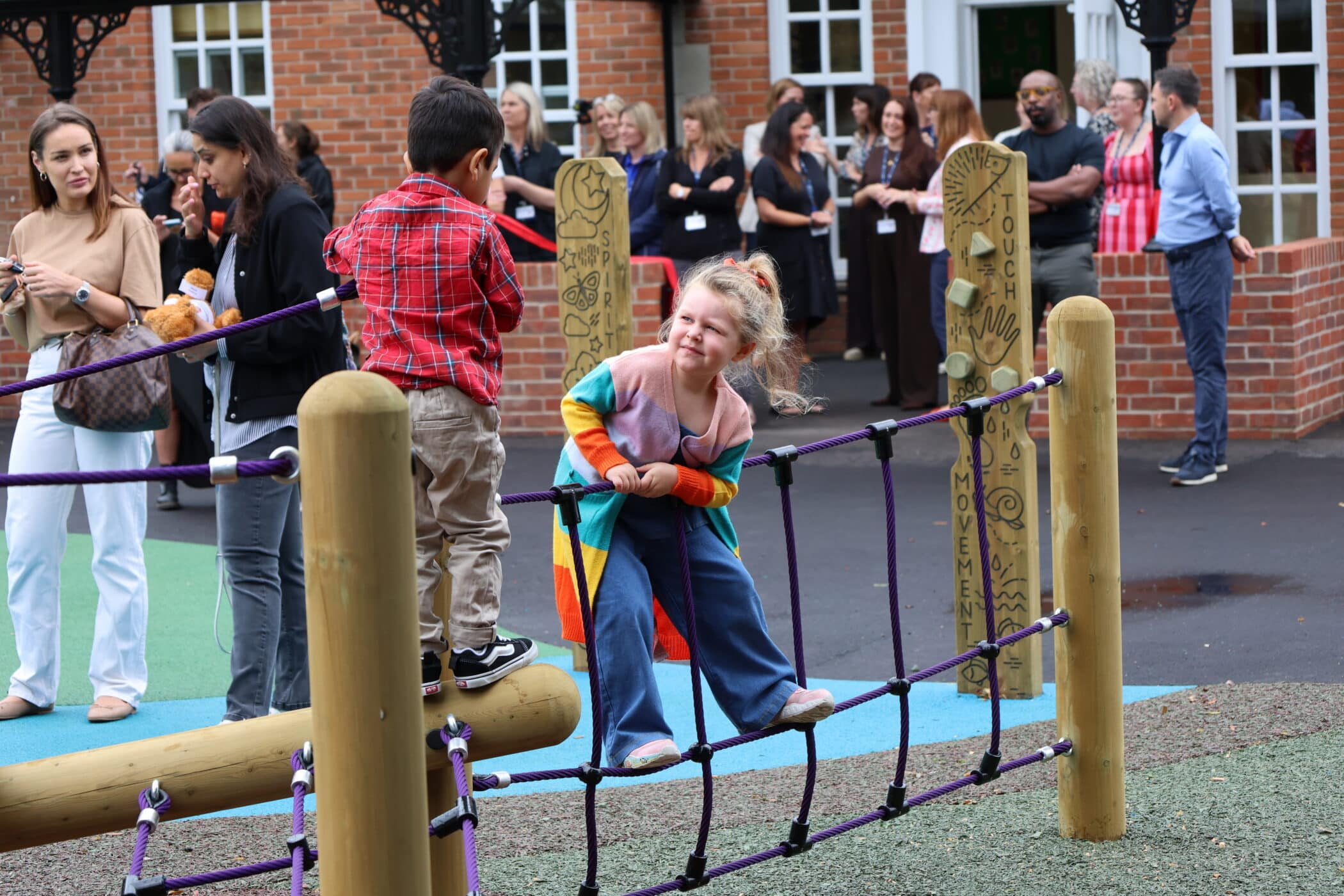 Two children play on a rope bridge at a playground while adults stand and watch in the background near a brick building.