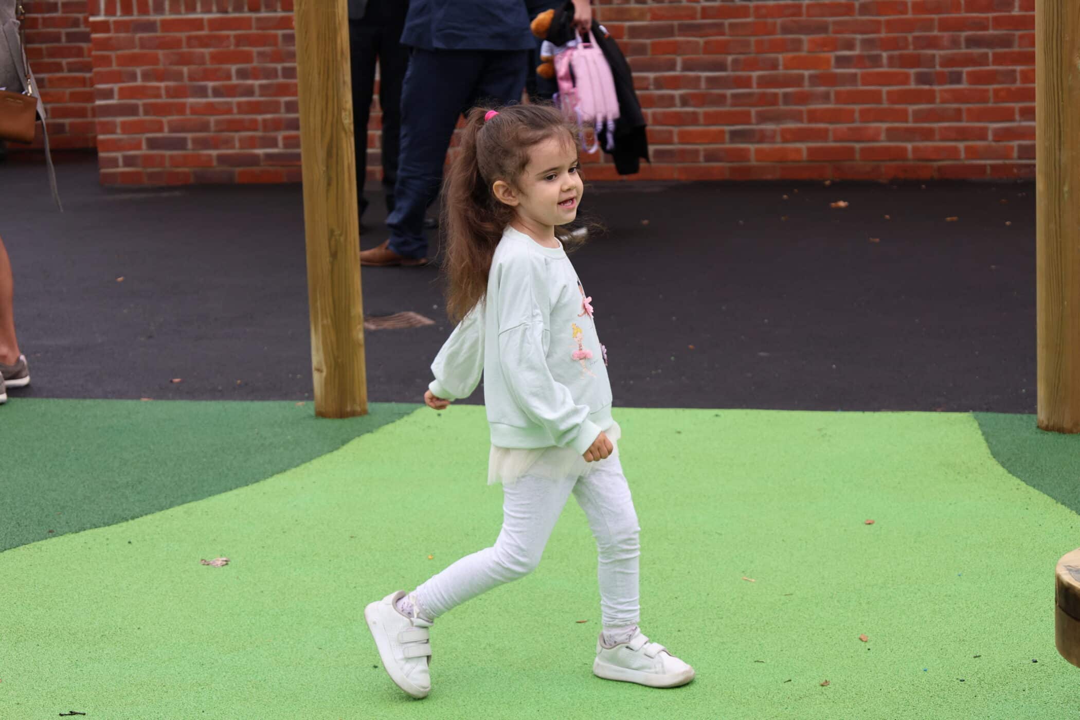 A young girl with long brown hair, wearing a light green jumper and white trousers, walks on a playground with green flooring and brick walls in the background.