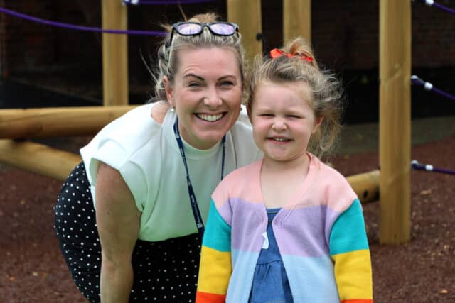 A smiling woman and young girl pose together outdoors by a wooden play structure. The woman wears a white top and polka dot skirt; the girl wears a colourful striped jumper and has her hair in a ponytail.