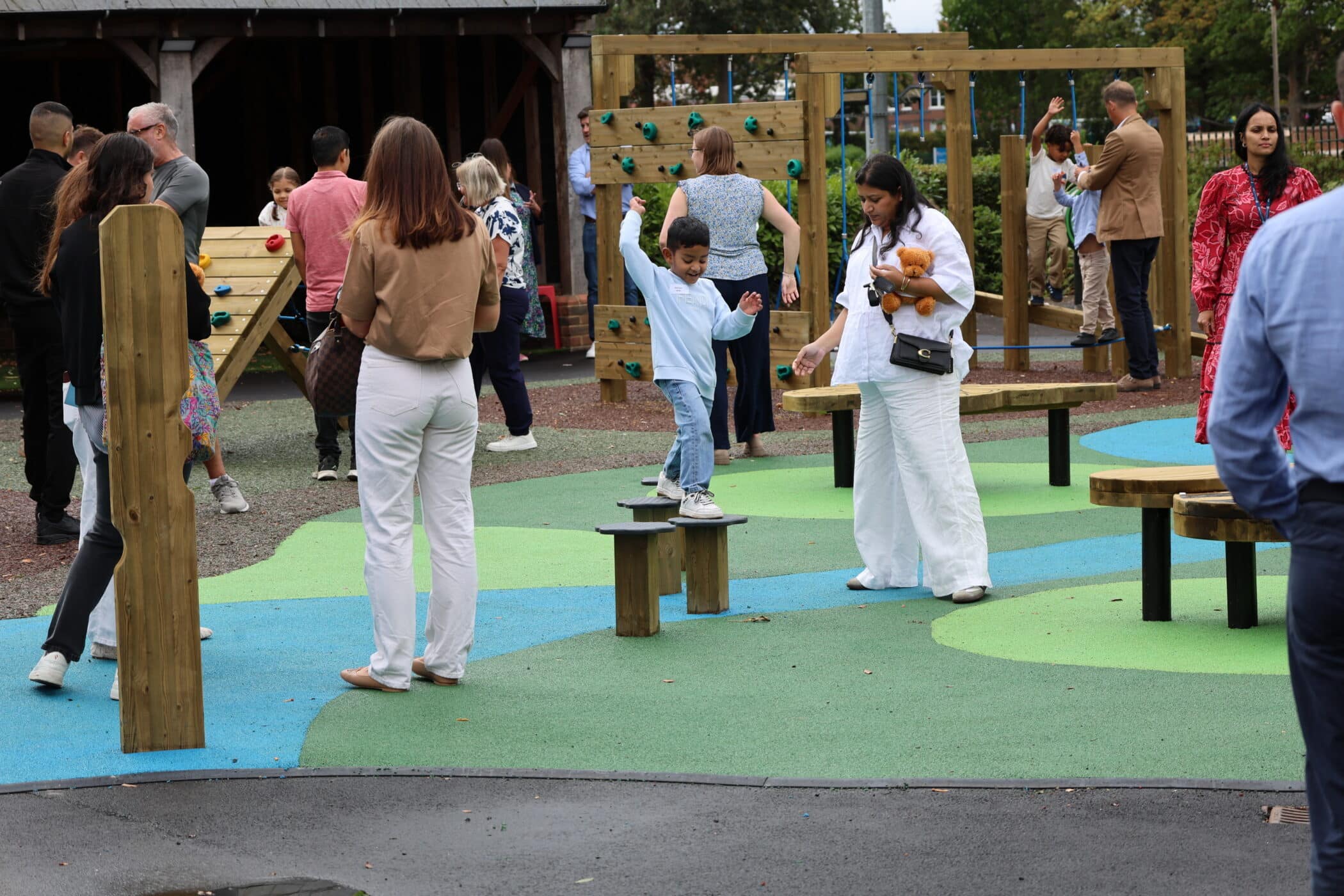 Children and adults gather at an outdoor play area with colourful flooring. A boy balances on a bench whilst others converse and supervise. The scene is lively and social.