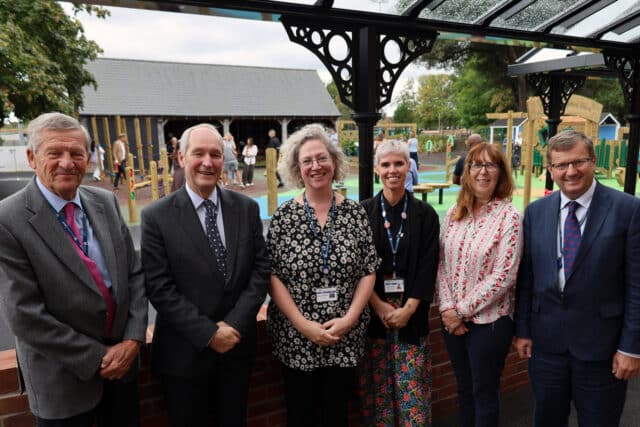 Six adults, four men and two women, stand smiling under a canopy in front of a school playground, dressed in business or smart casual attire, with colourful playground equipment visible in the background.