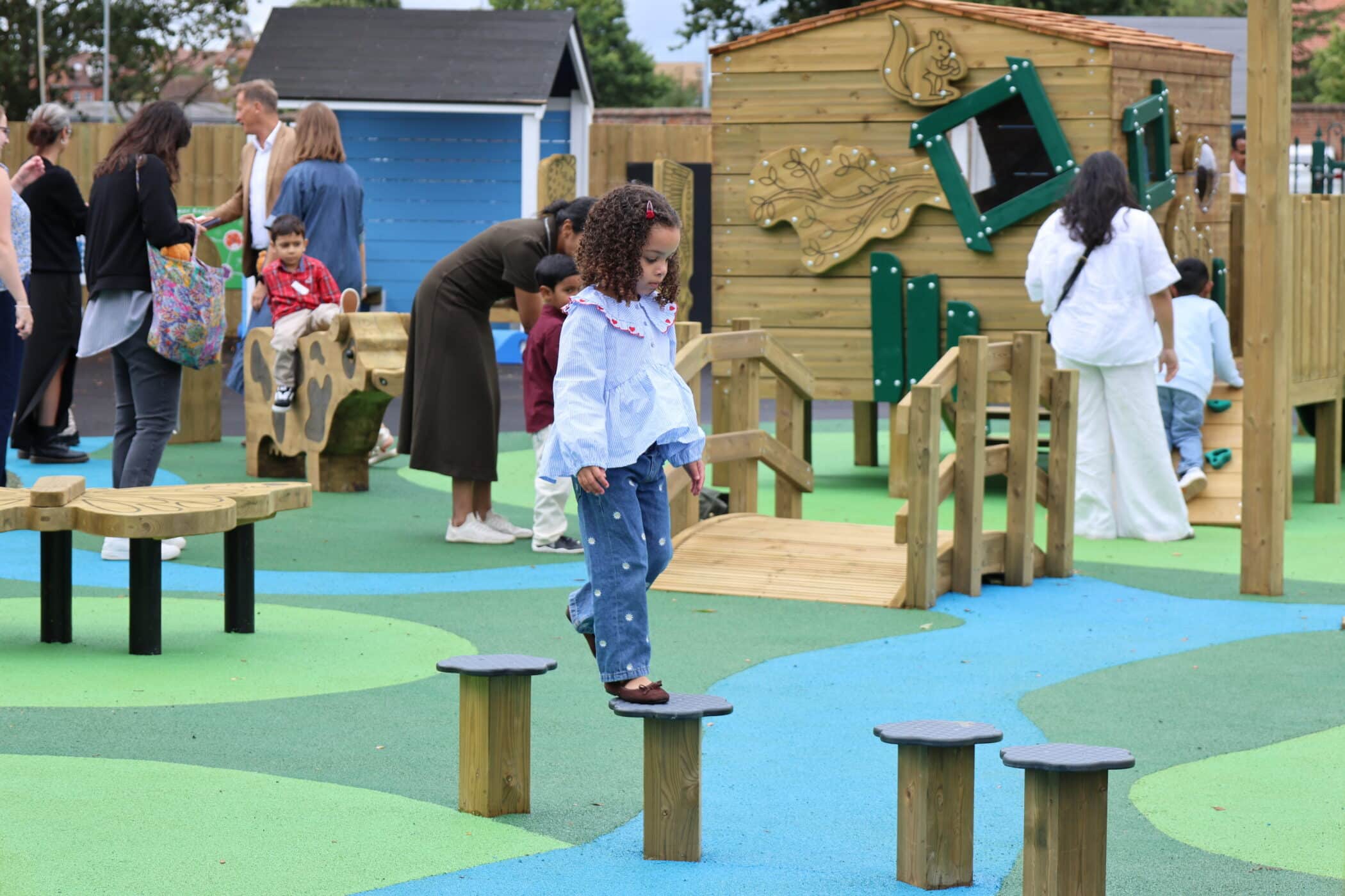 Children and adults gather at a colourful playground. A young girl carefully balances on stepping stones, whilst others explore climbing frames and interact nearby.