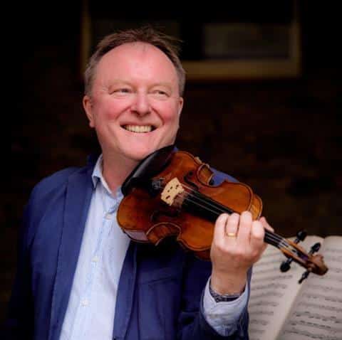 A smiling man in a blue jacket holds a violin and bow, standing in front of a music stand with sheet music.