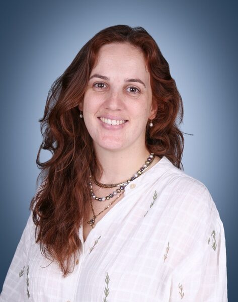 A woman with long brown hair smiles at the camera. She wears a white shirt with a subtle pattern and beaded necklaces, set against a blue gradient background.