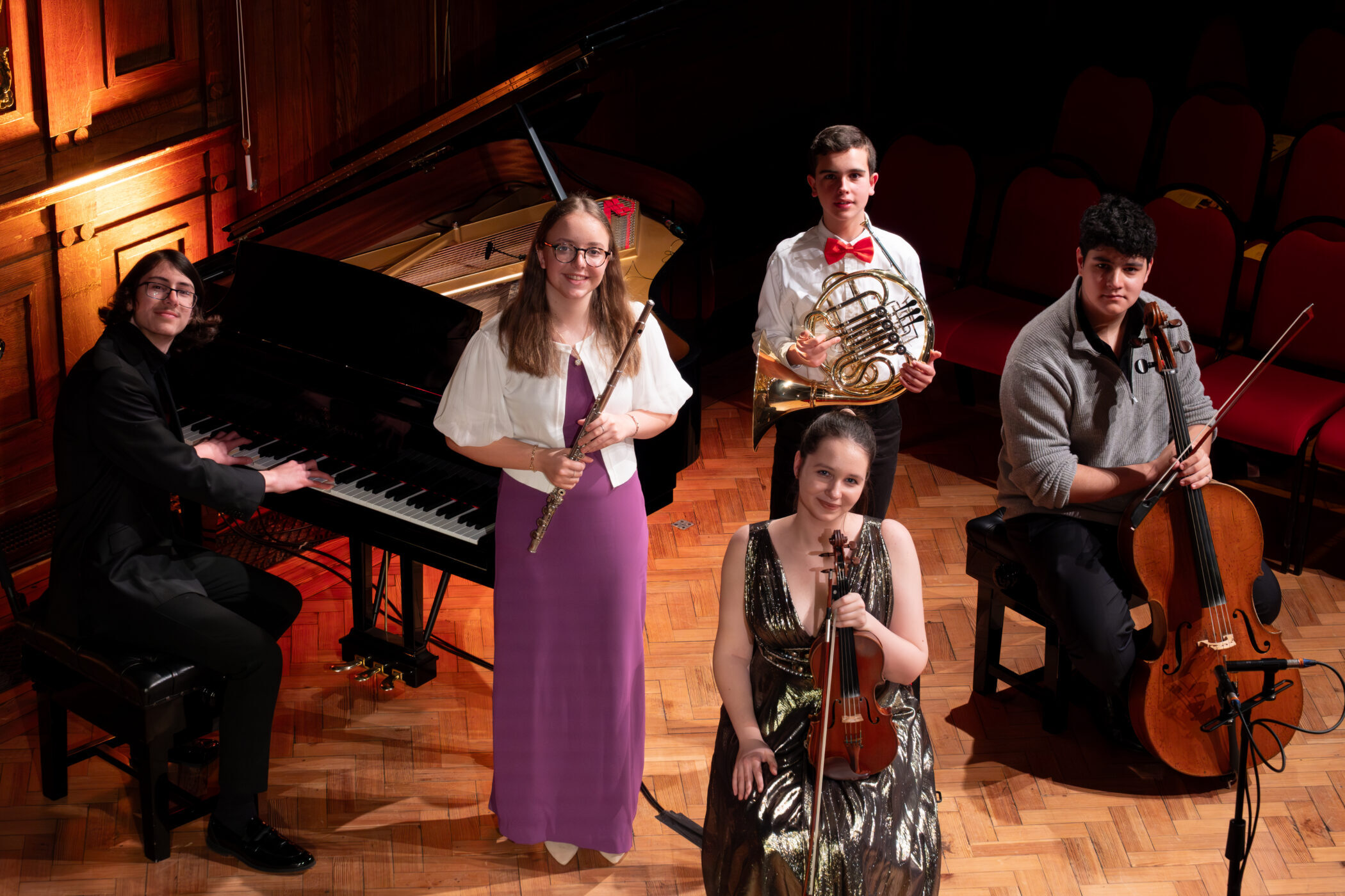 Five musicians pose on a wooden stage with their instruments, piano, flute, French horn, violin, and cello, in celebration of the Brentwood International Music Competition. Four are seated, one stands, and they all smile at the camera under warm lighting.
