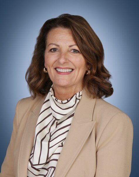 A woman with shoulder-length brown hair, wearing a beige jacket and a white blouse with black stripes, smiles at the camera against a blue gradient background.
