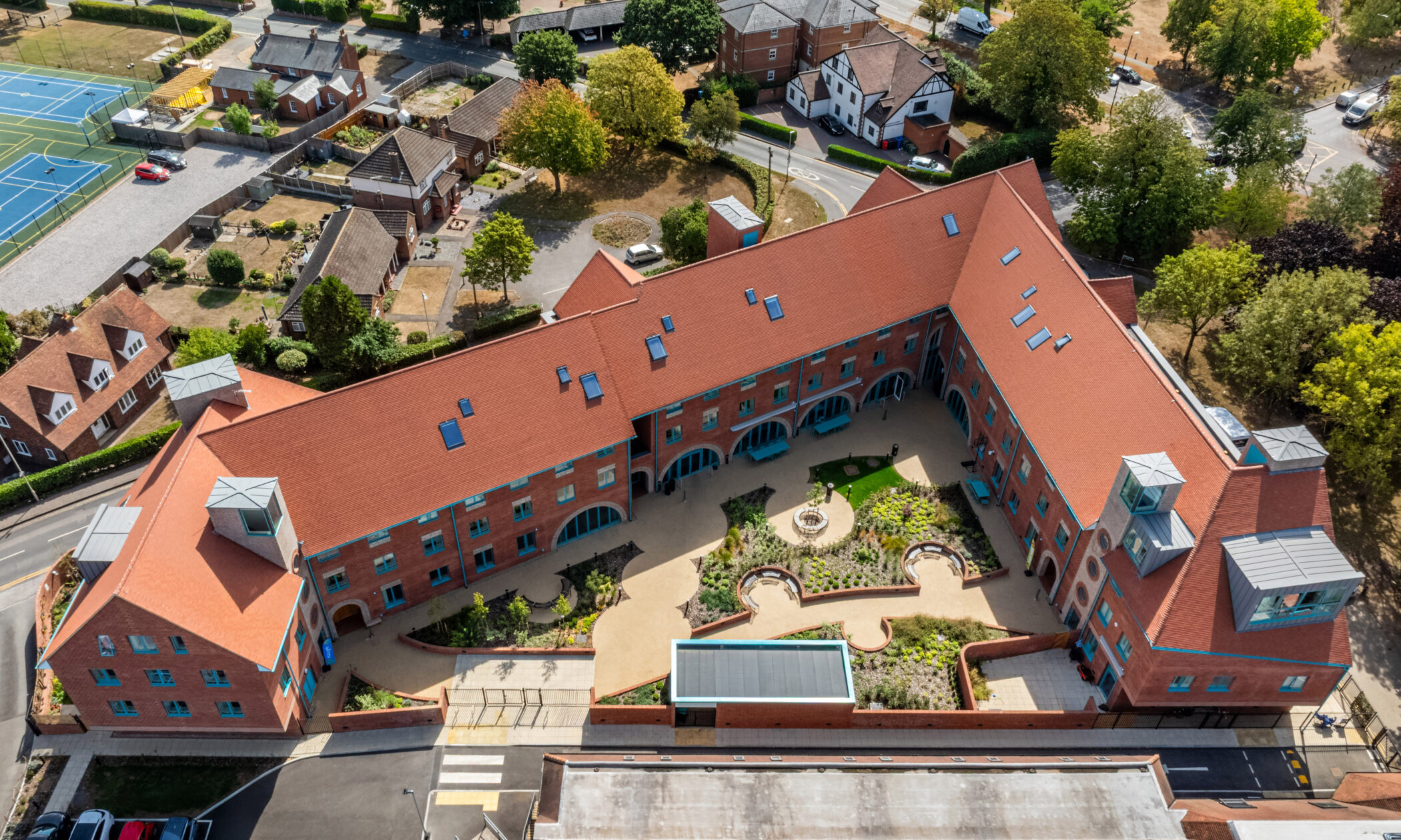 Aerial view of a large, red-roofed, L-shaped building surrounding a landscaped central courtyard, with nearby houses, trees, tennis courts, and parking areas visible, all highlighting the extensive facilities available on the property.