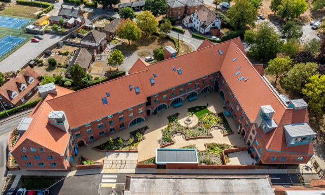 Aerial view of a large, red-roofed, L-shaped building surrounding a landscaped central courtyard, with nearby houses, trees, tennis courts, and parking areas visible, all highlighting the extensive facilities available on the property.