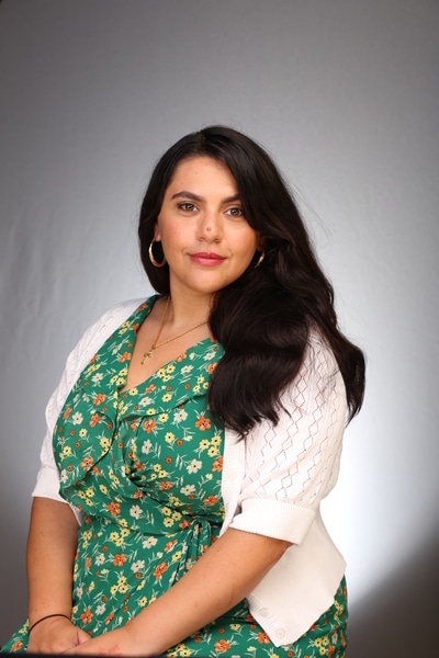 Woman with long dark hair in a green floral dress and white jumper sits in front of a plain grey background, looking calmly at the camera.