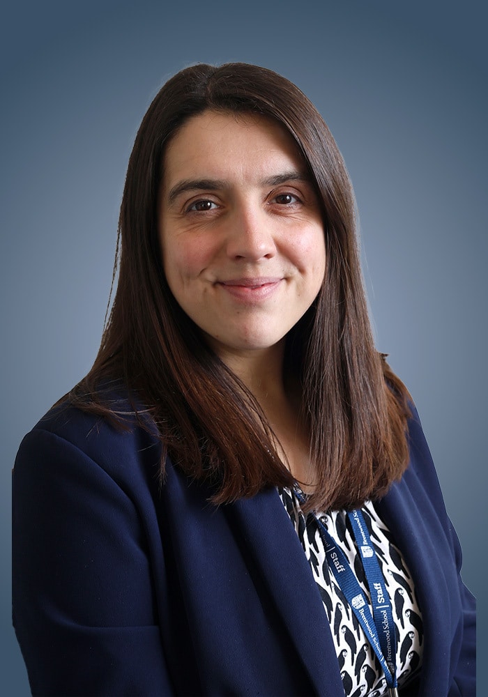 A woman with straight brown hair wearing a navy blazer and patterned top smiles at the camera. She has a staff lanyard around her neck, indicating her pastoral role, and the background is a soft blue gradient.