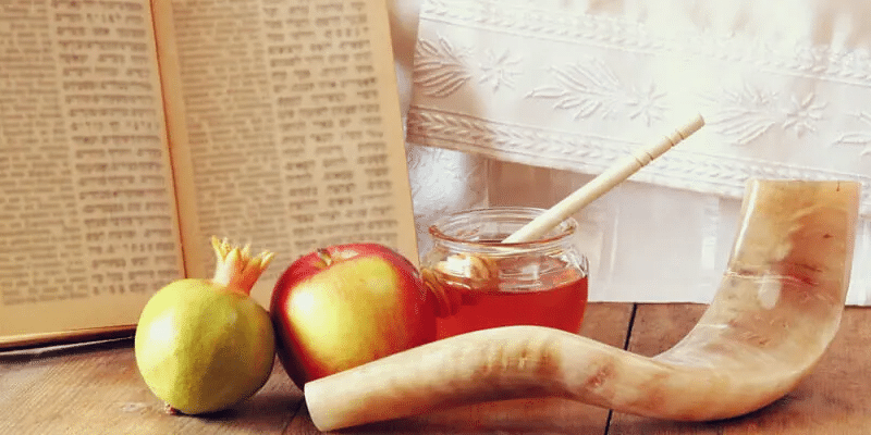 Open book, apple, pomegranate, honey jar with drizzler, and a shofar on a wooden table, with a white embroidered cloth in the background.