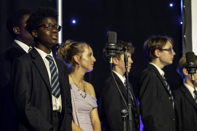 A group of students in suits and a woman in a purple dress stand in front of microphones on stage, under stage lights.